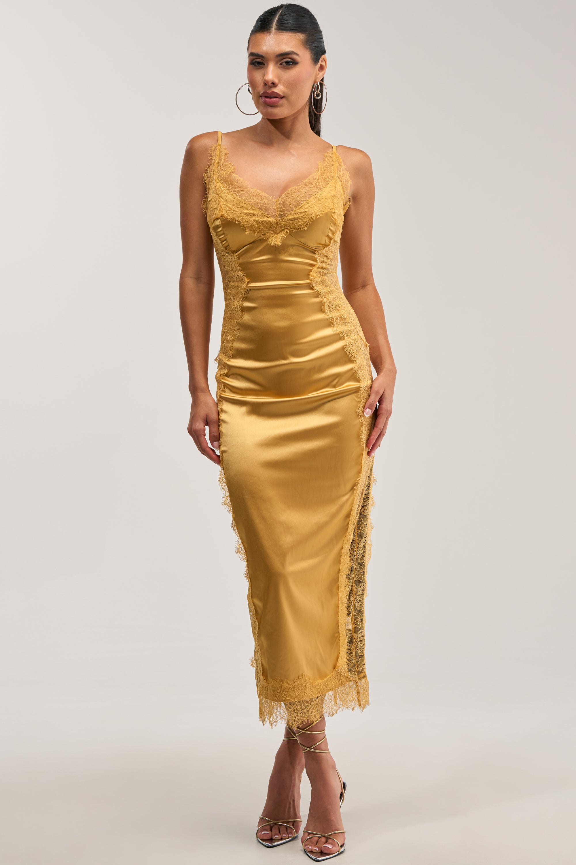 Wearing the ESSENTIAL LACE TRIM SATIN SLEEVELESS MIDI DRESS IN GOLD—with its delicate lace accents and sleek spaghetti straps—this woman pairs the effortlessly elegant cocktail look with strappy heels, standing poised against a simple light background.