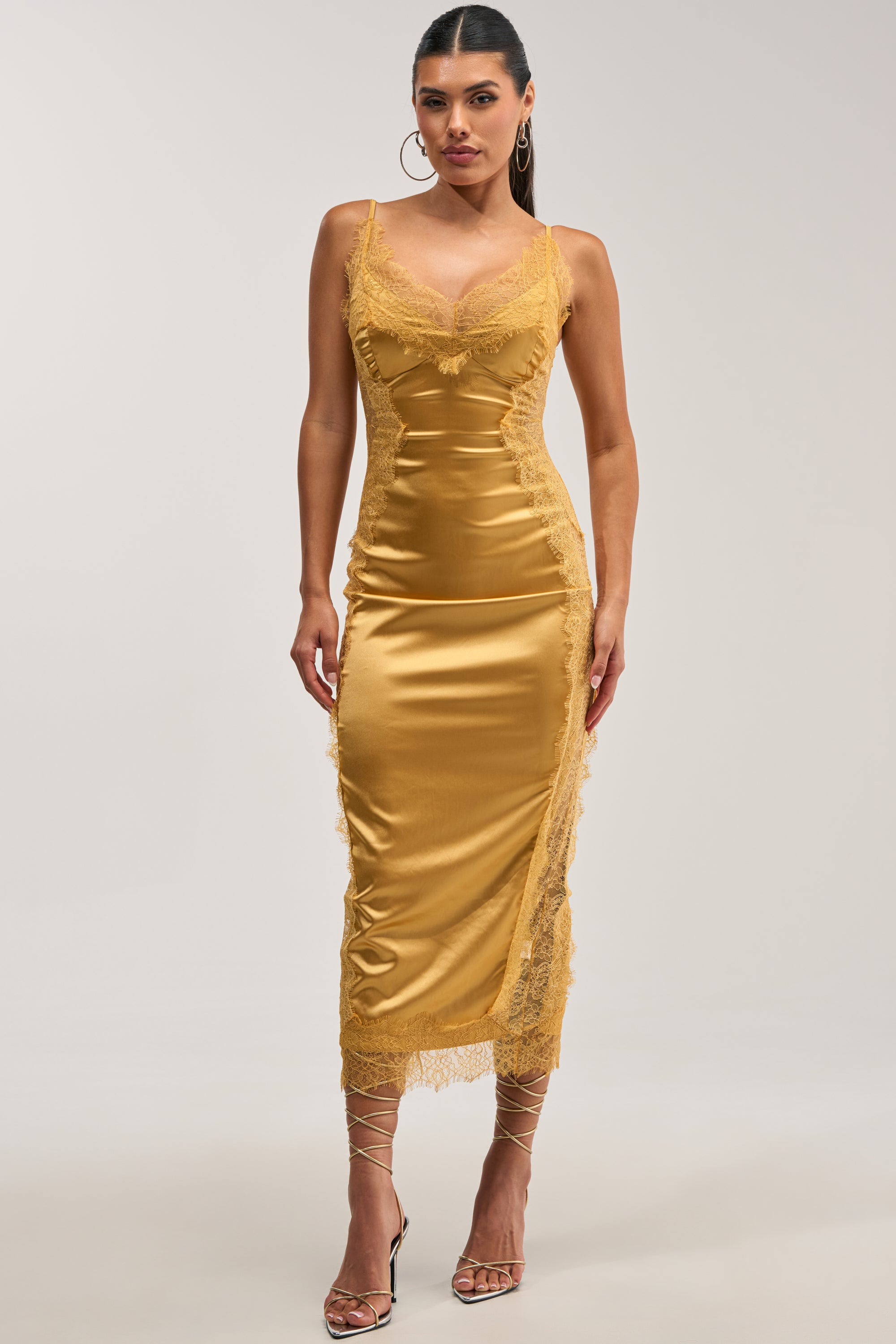 A woman with dark hair styled in a ponytail poses against a grey background, wearing the ESSENTIAL LACE TRIM SATIN SLEEVELESS MIDI DRESS IN GOLD, which features delicate lace trim at the neckline, sides, and hem. She completes her sophisticated evening cocktail look with gold lace-up high-heeled sandals and gold hoop earrings.