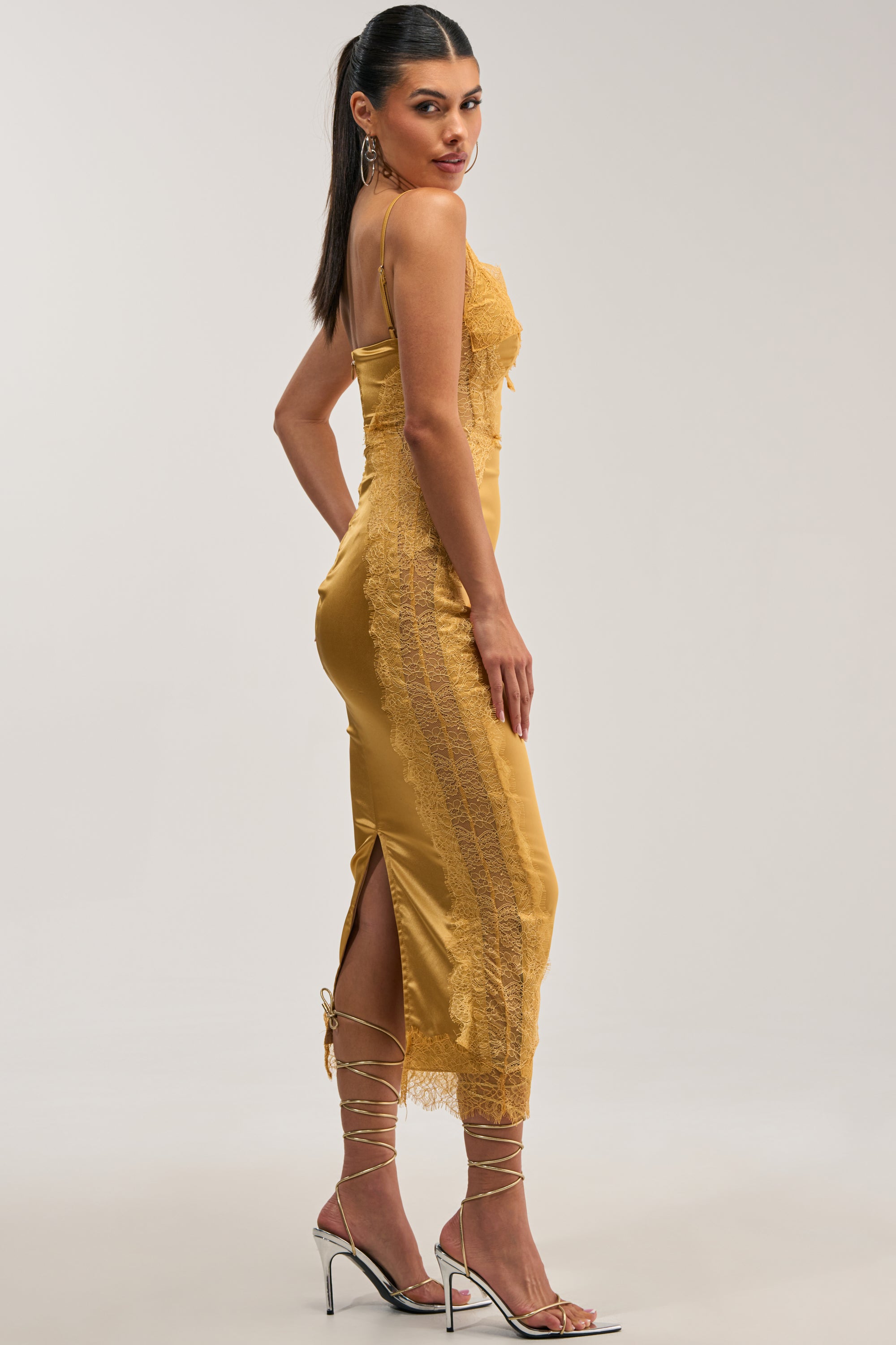 A model with a high ponytail wears the ESSENTIAL LACE TRIM SATIN SLEEVELESS MIDI DRESS IN GOLD, which features delicate lace panels along the bodice and sides. She pairs the dress with lace-up, strappy gold heels while posing against a neutral studio background.