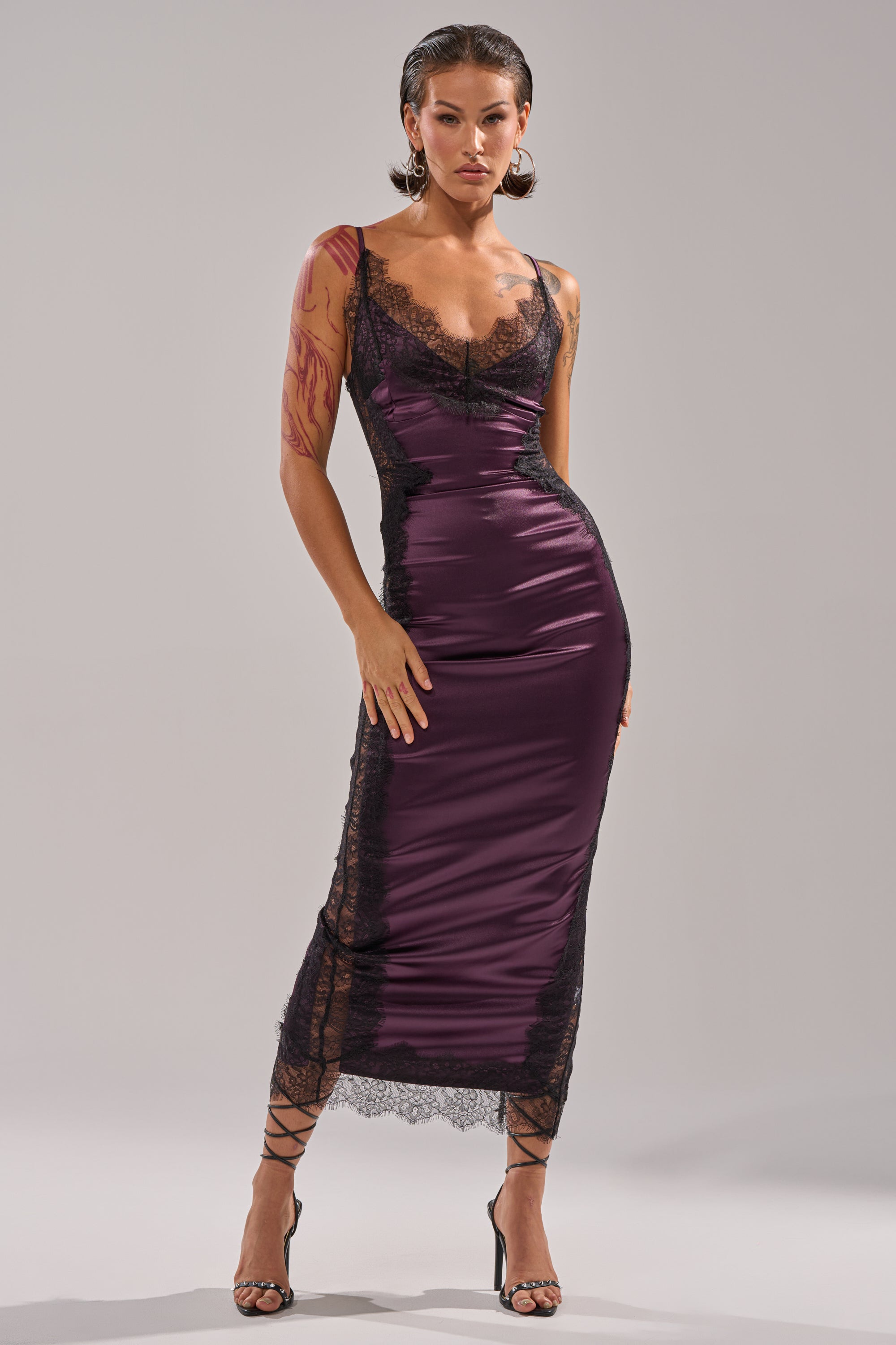 A woman stands confidently in the ESSENTIAL LACE TRIM SATIN SLEEVELESS MIDI DRESS—fitted, sleeveless, with black lace trim—paired with black strappy heels and slicked-back hair against a plain light gray background for a chic look.