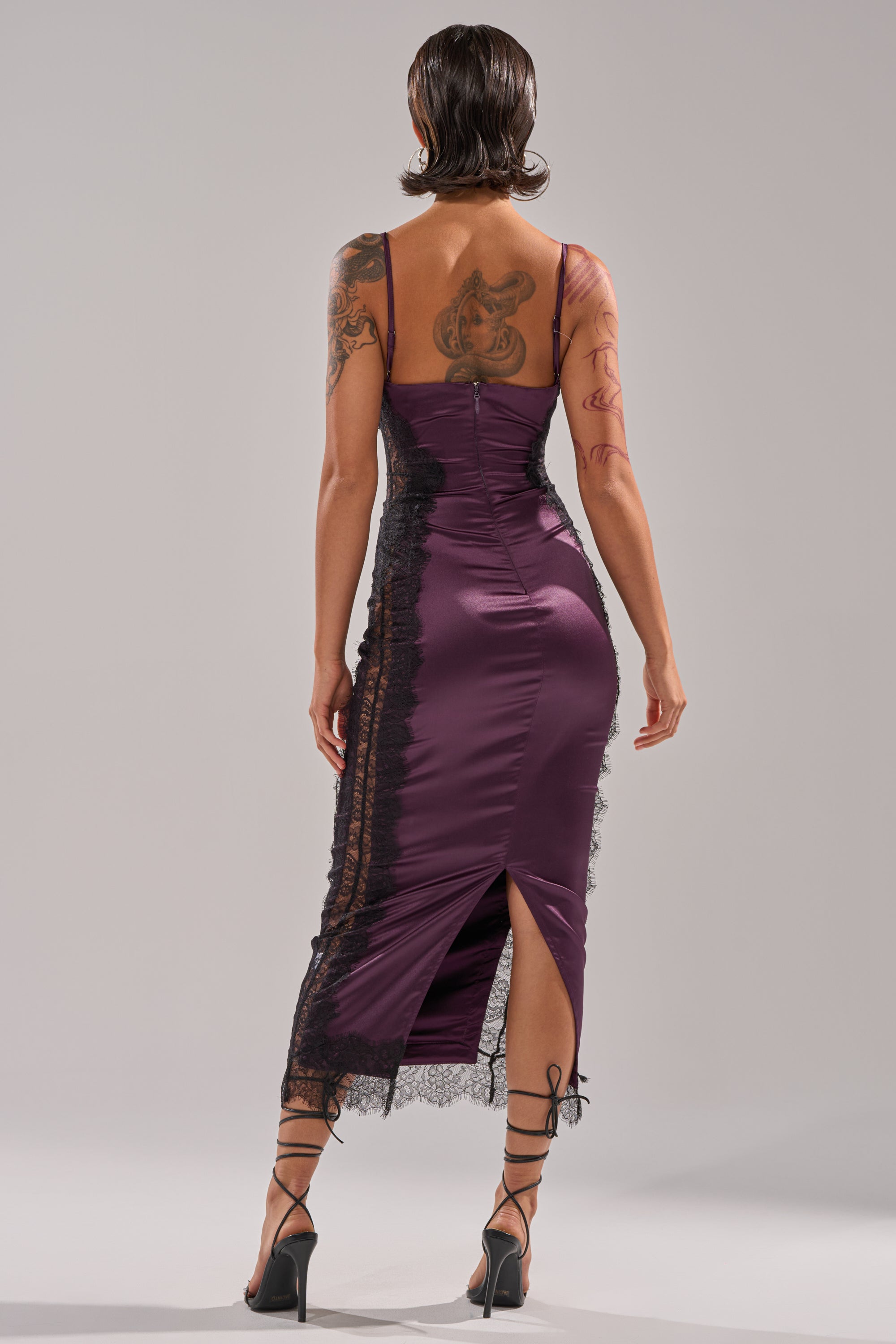 A person with short dark hair, visible tattoos, and strappy high heels faces away wearing the ESSENTIAL LACE TRIM SATIN SLEEVELESS MIDI DRESS in purple, featuring a fitted silhouette and lace trim.