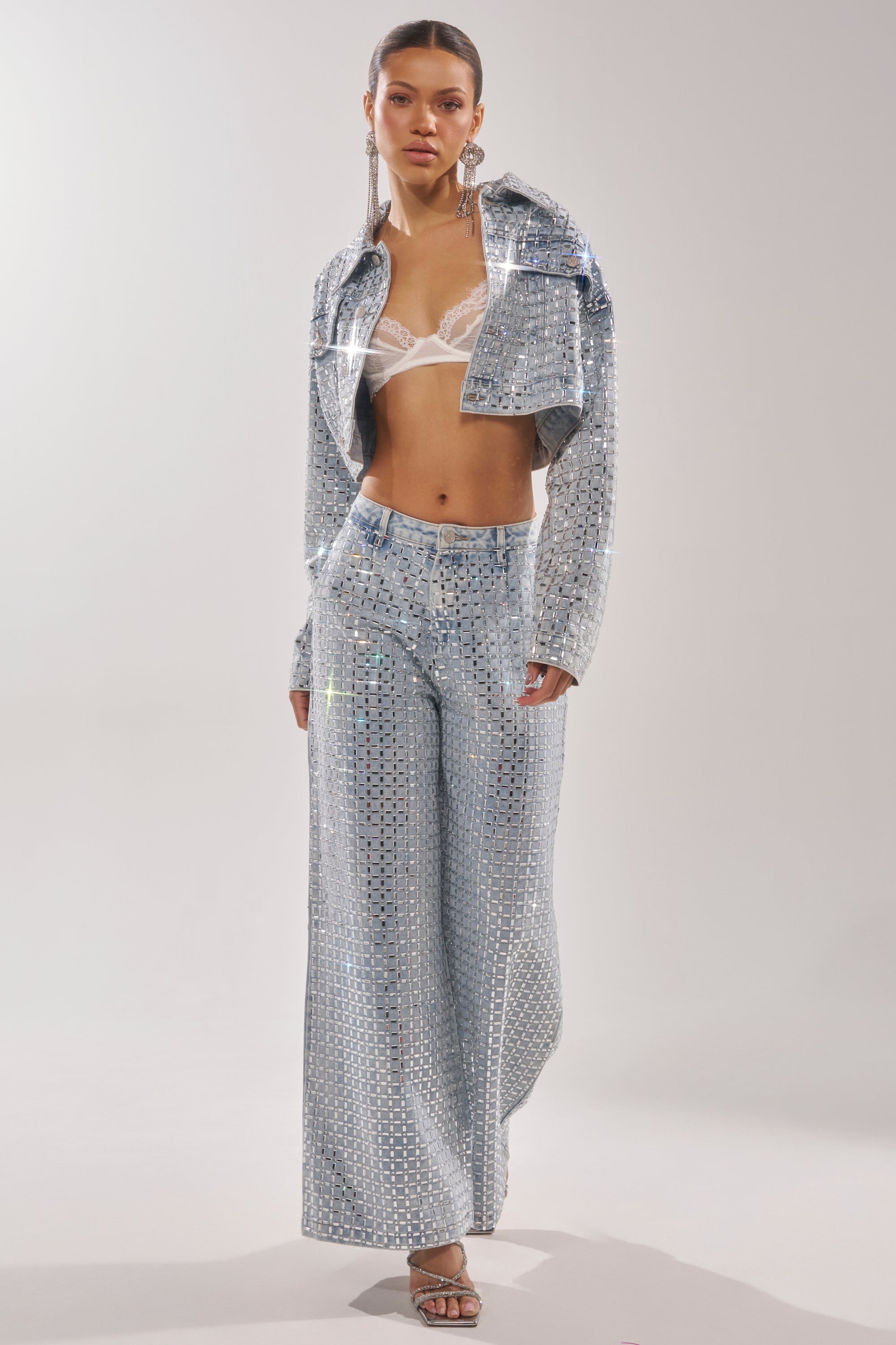 A woman showcases the PICK N CHOOSE RHINESTONE DENIM JACKET, styled over a sheer white lace bralette with hoop earrings and heeled sandals, posed against a plain light background. 10