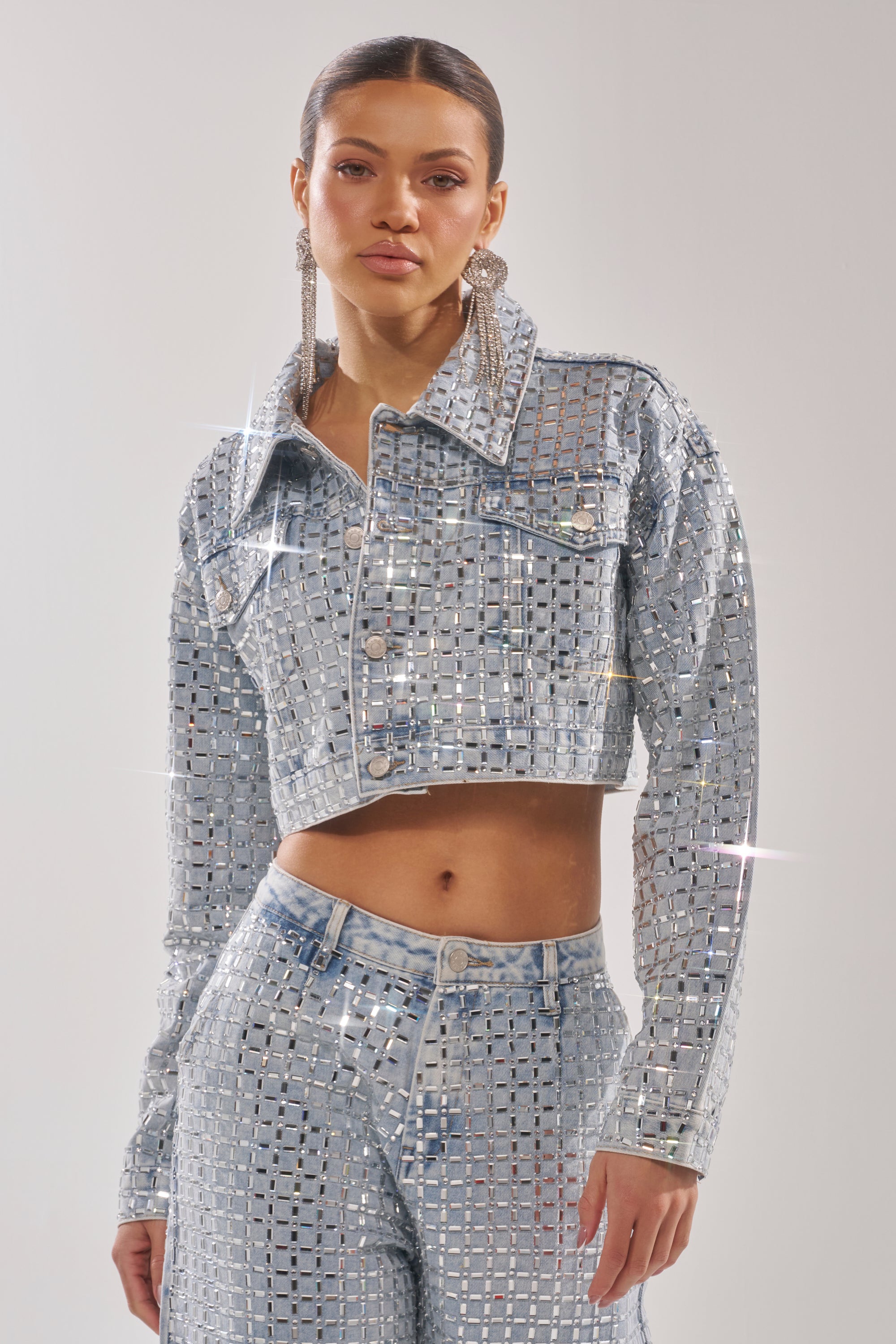 A woman wears the PICK N CHOOSE RHINESTONE DENIM JACKET, cropped and sparkling, paired with matching high-waisted pants and statement earrings. The outfit catches the light for a shimmering effect. 1