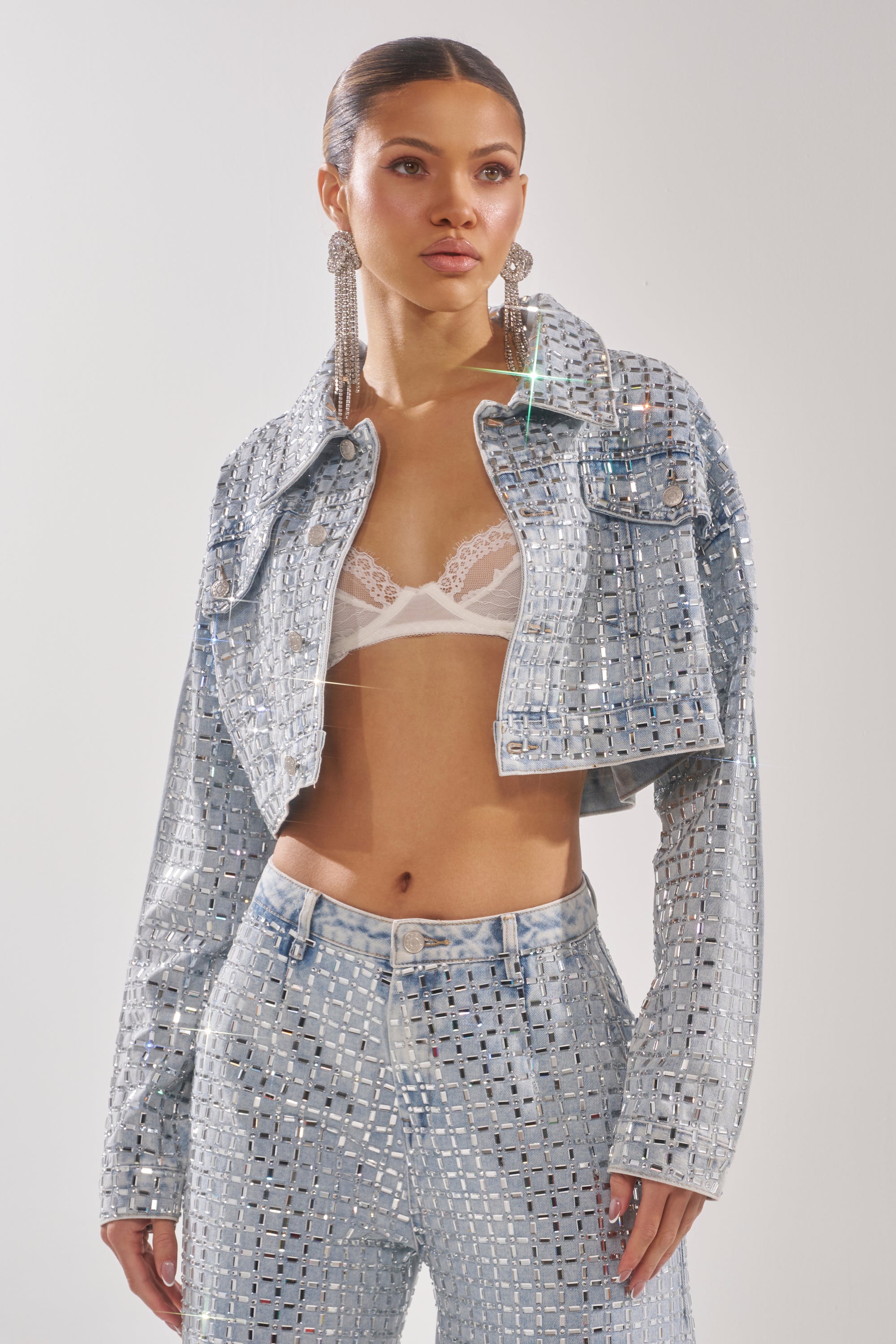 A woman wears the PICK N CHOOSE RHINESTONE DENIM JACKET and matching pants over a white lace bralette, accessorized with long earrings and her hair pulled back. The background is plain and light-colored. 2