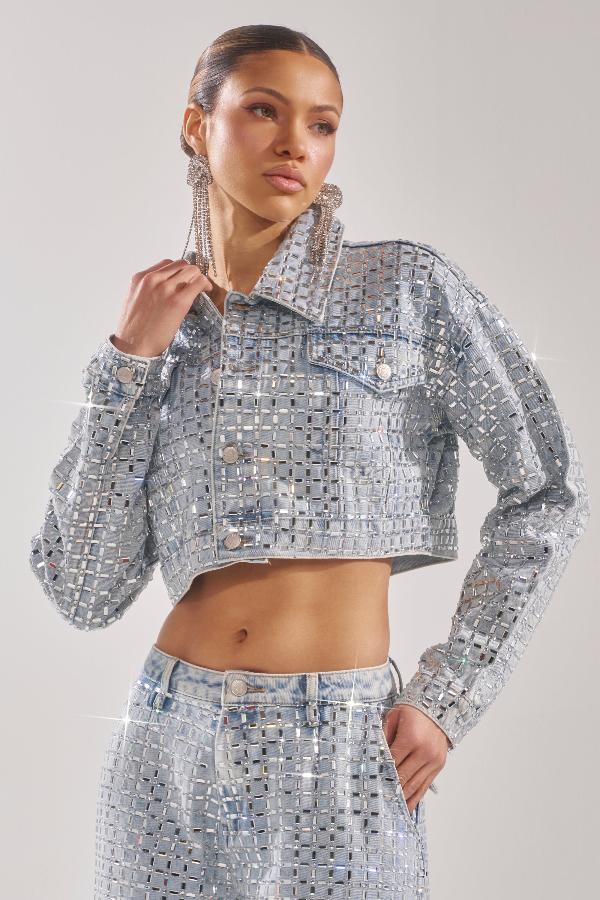 A woman wears the PICK N CHOOSE RHINESTONE DENIM JACKET with matching high-waisted pants featuring square cutouts and mirror embellishments. She accessorizes with large dangling earrings and has her hair pulled back. 4
