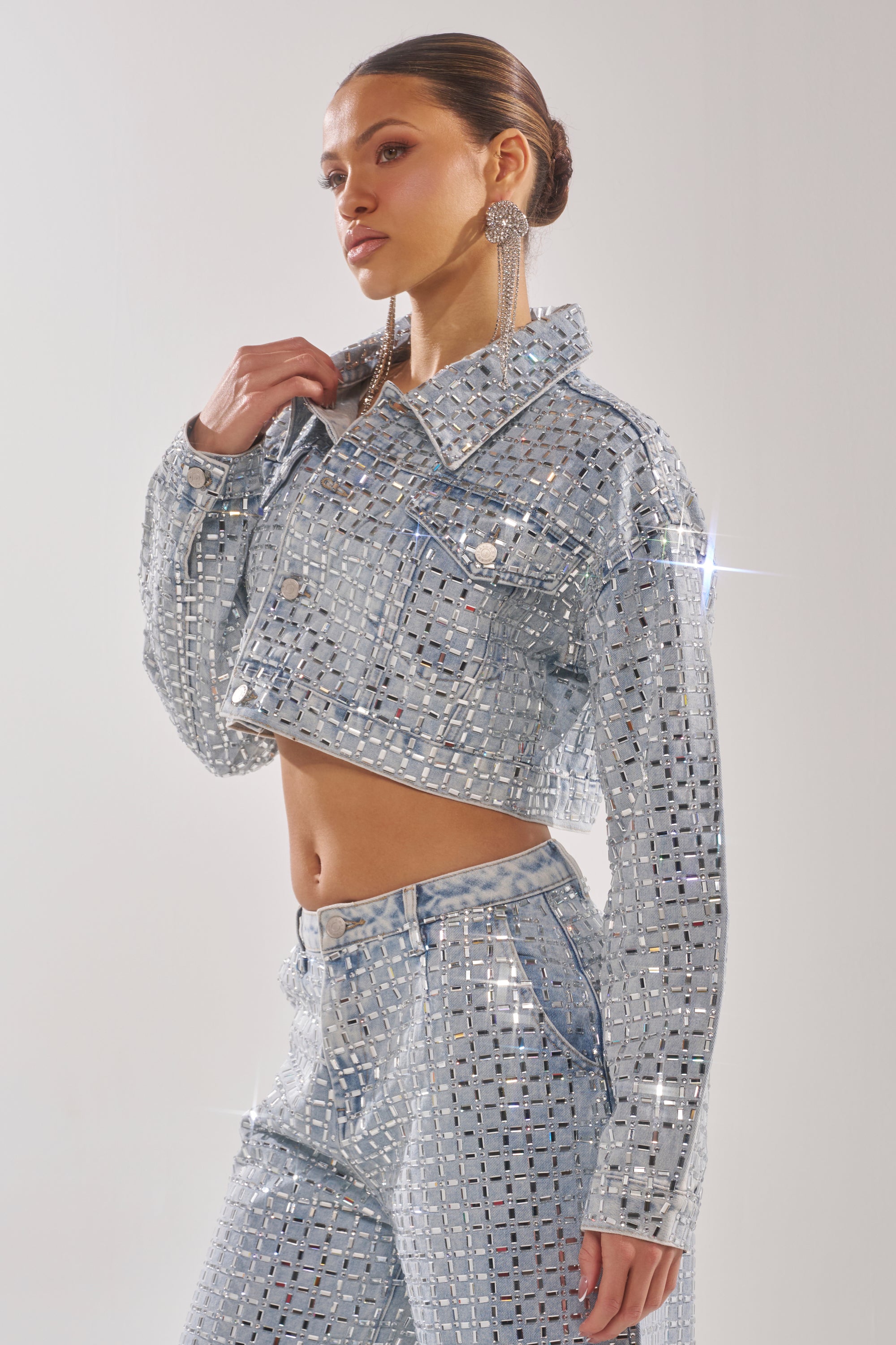 A woman poses in the PICK N CHOOSE RHINESTONE DENIM JACKET and matching high-waisted pants, accessorized with statement earrings and slicked-back hair, shimmering against a plain light background. 5