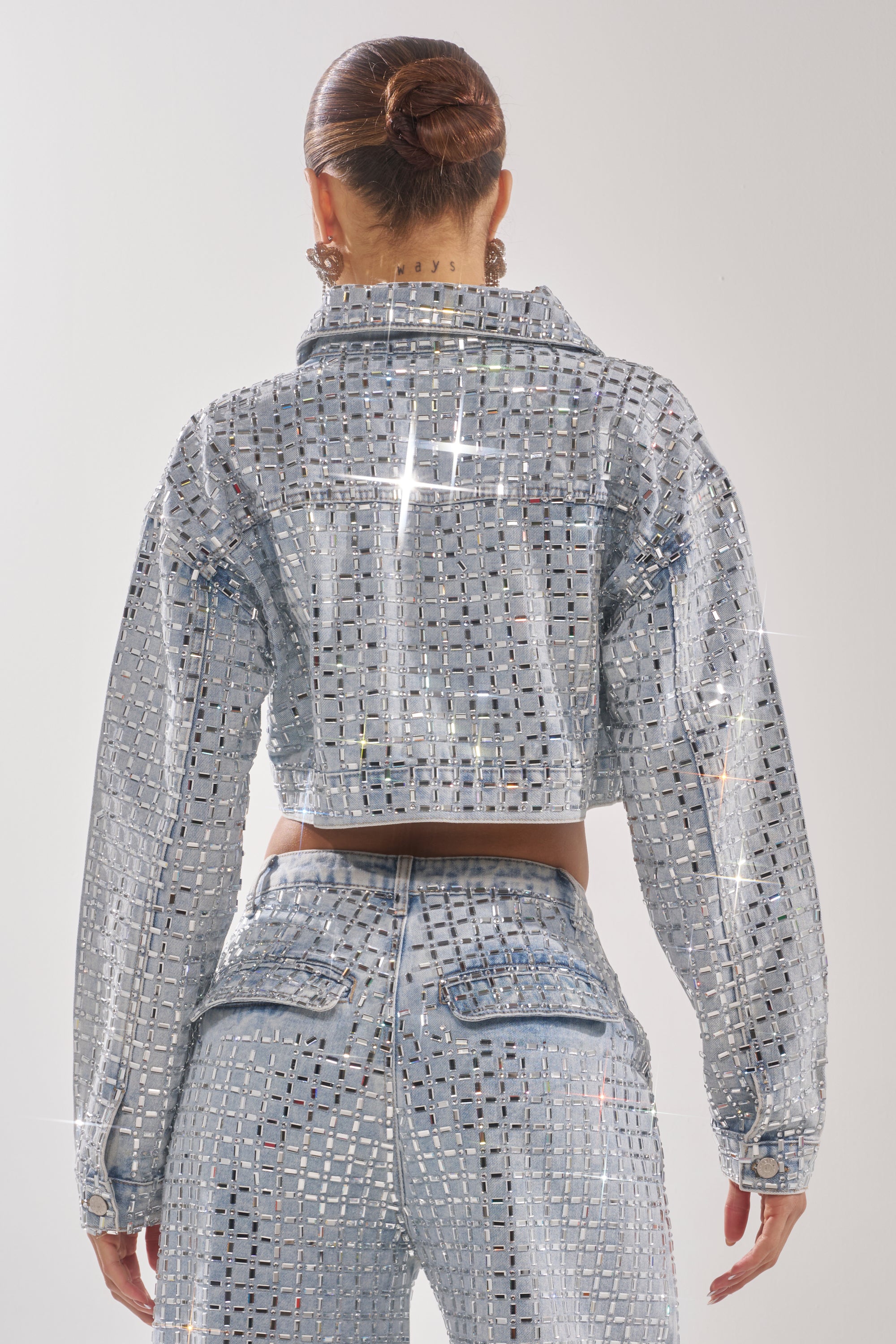 A person with hair in a sleek bun models the PICK N CHOOSE RHINESTONE DENIM JACKET, sparkling and silver-embellished, paired with matching high-waisted pants, seen from behind against a plain light background. 6