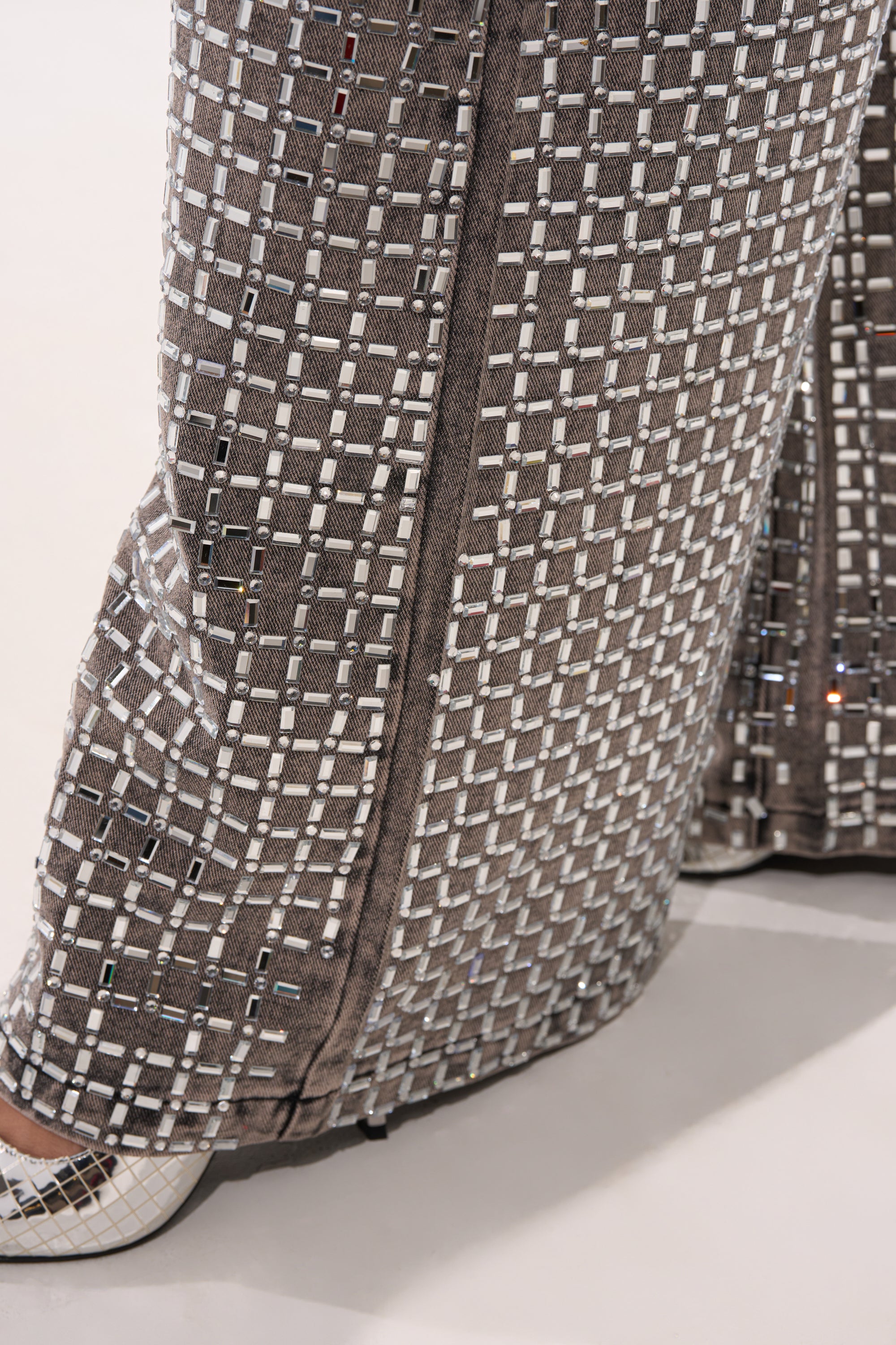 A close-up of a person's lower legs showcases the PICK N CHOOSE RHINESTONE WIDE LEG DENIM IN GREY with a geometric silver and white grid pattern, paired with white shoes against a plain, light background—ideal for chic evening wear. 9