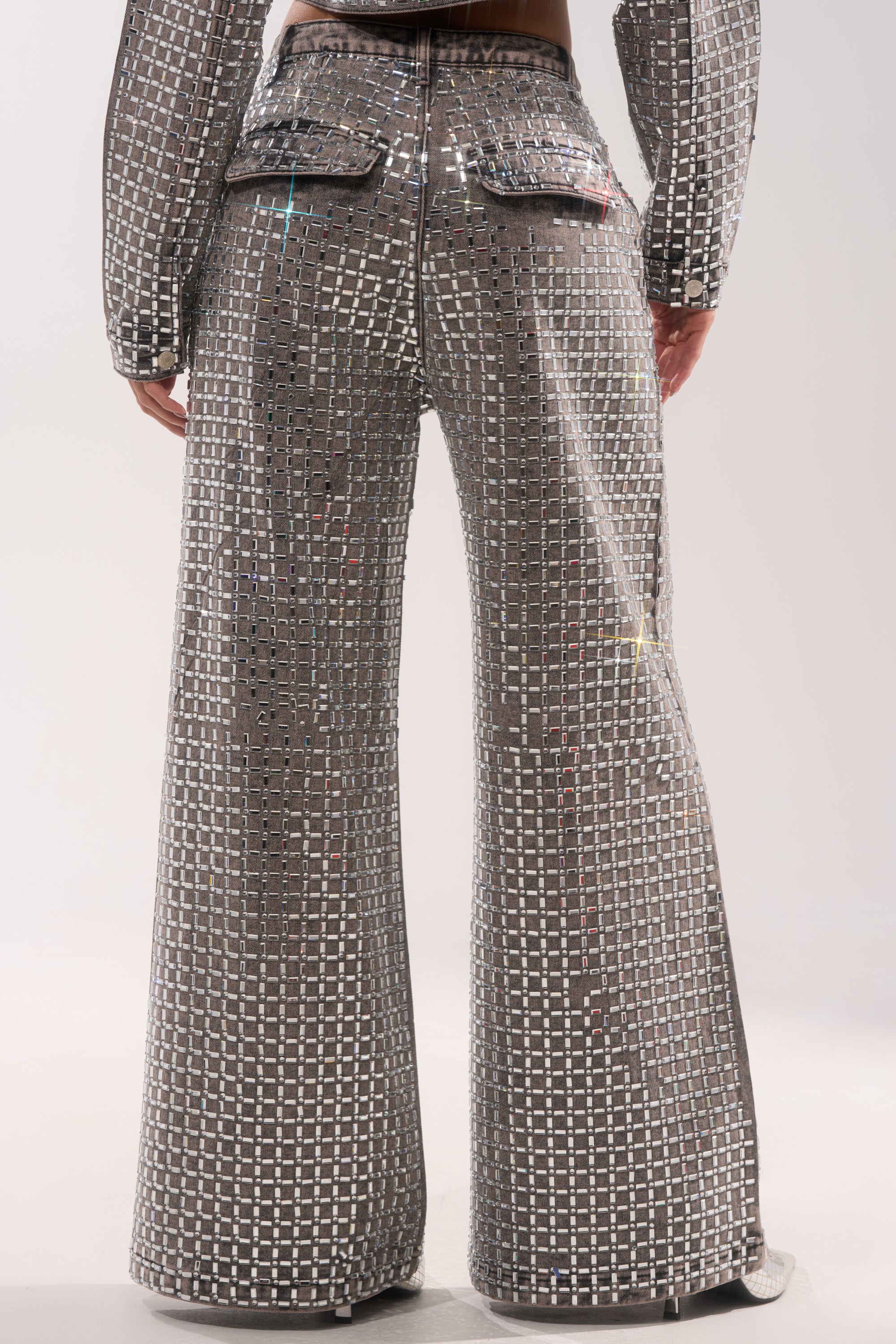 A person wearing the PICK N CHOOSE RHINESTONE WIDE LEG DENIM IN GREY—shiny, silver, wide-leg jeans with a grid of reflective squares—shown from the back against a plain light background, ideal for bold evening style. 6