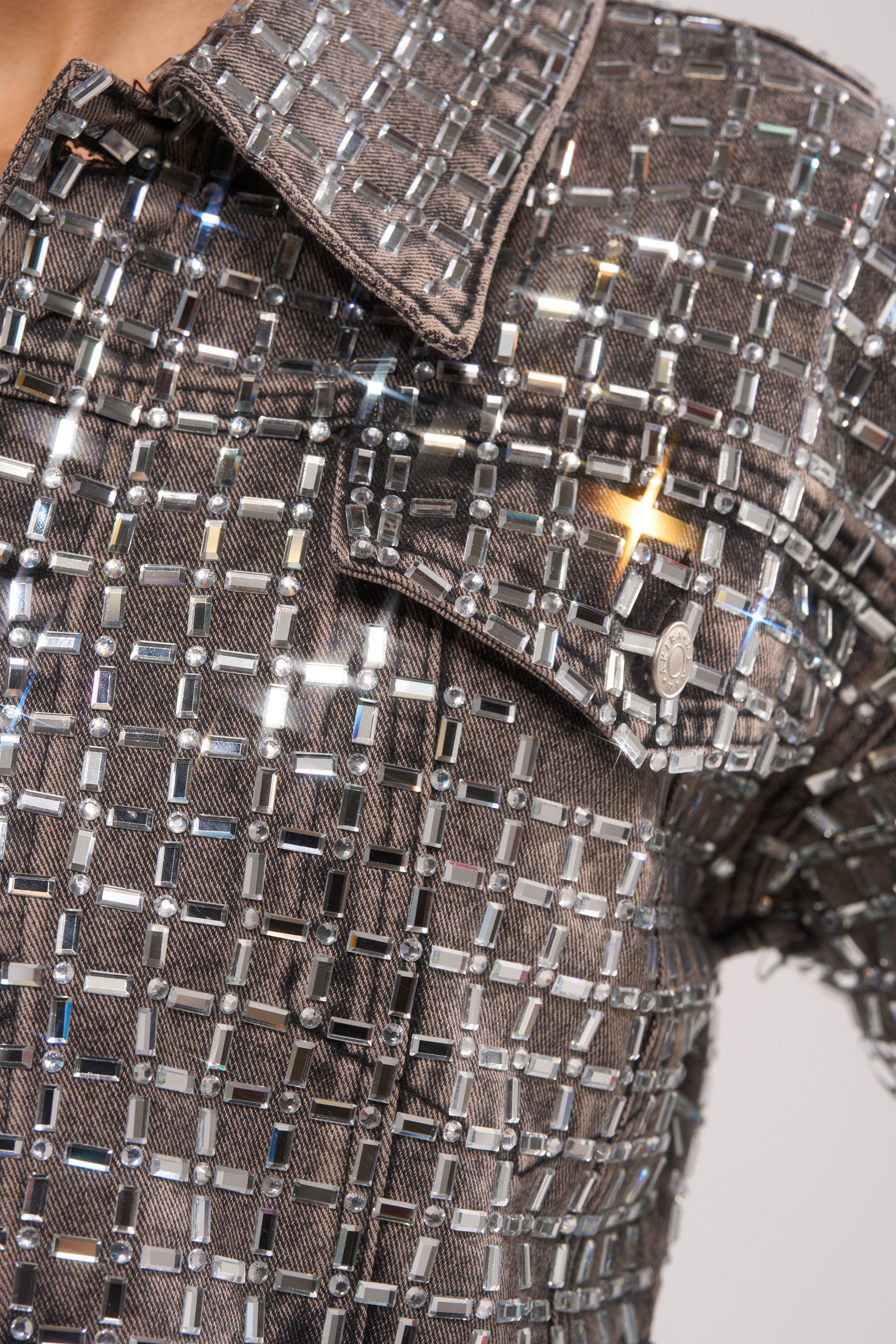 Close-up of a person wearing the PICK N CHOOSE RHINESTONE DENIM JACKET IN GREY, adorned with reflective rhinestones in a grid pattern for a statement street style look. Features include a collar and buttoned chest pocket. 10