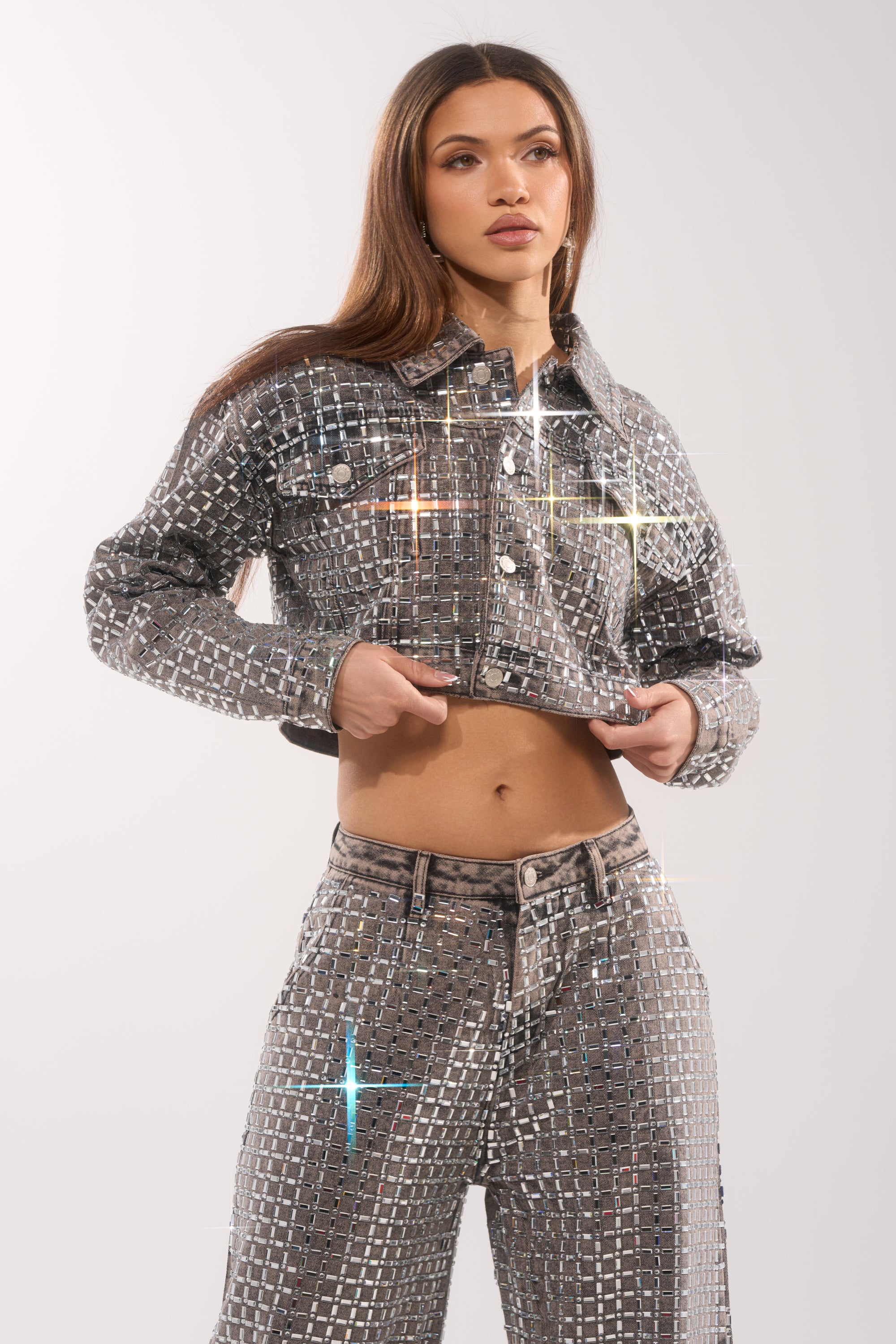 A woman poses in the PICK N CHOOSE RHINESTONE DENIM JACKET IN GREY, its silver, grid-patterned fabric sparkling with edited highlights as she lifts the jacket to reveal her midriff. 1
