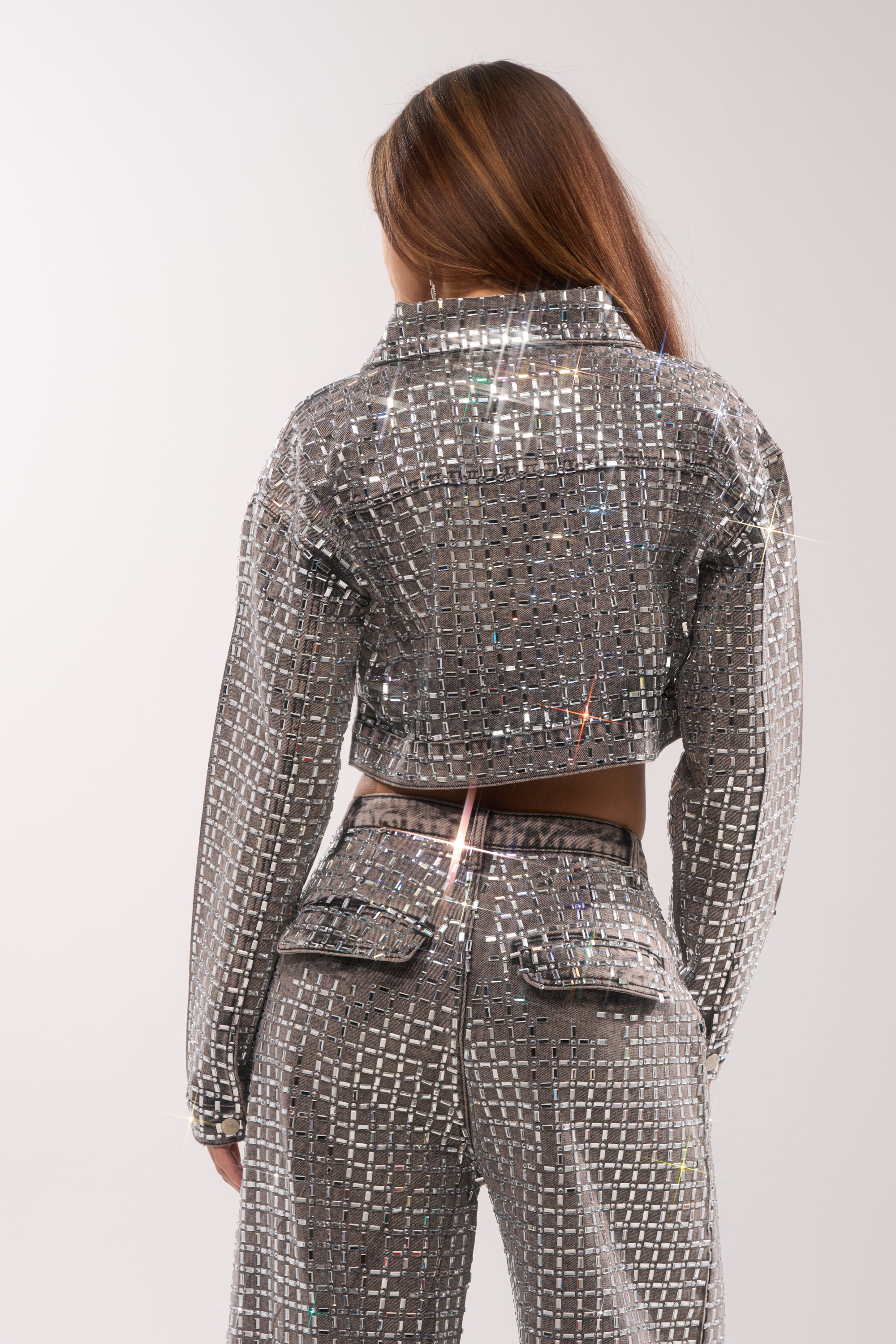 A person with long hair, seen from behind, wears the PICK N CHOOSE RHINESTONE DENIM JACKET IN GREY—a shiny, silver, reflective cropped jacket—paired with high-waisted pants embellished with small mirror-like squares. 6