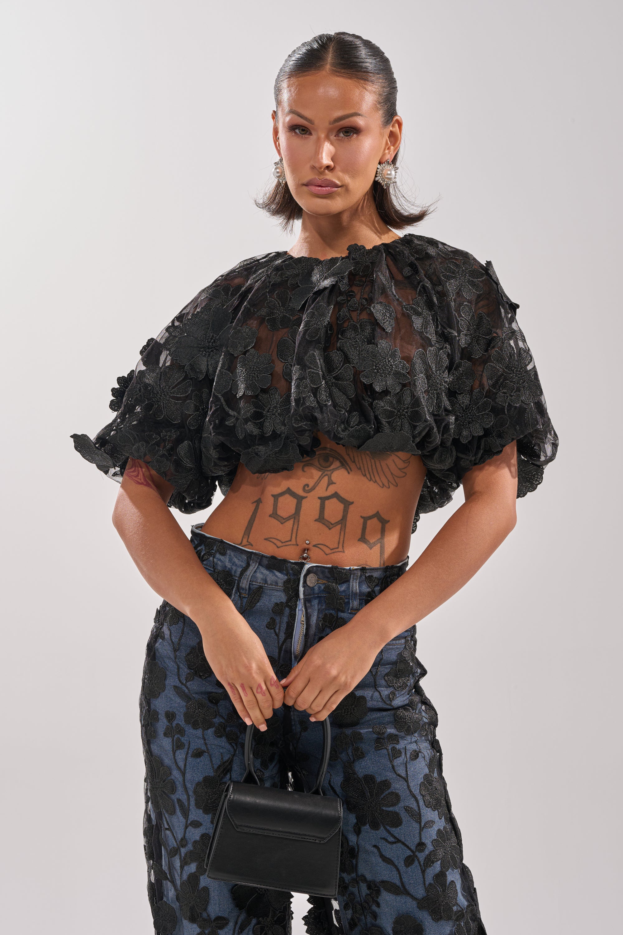 A woman with slicked-back hair wears the DONT GIVE UP LACE BLOUSE, blue floral pants, and holds a small black handbag. She displays a large "1999" tattoo on her torso and stands against a plain light background. 0