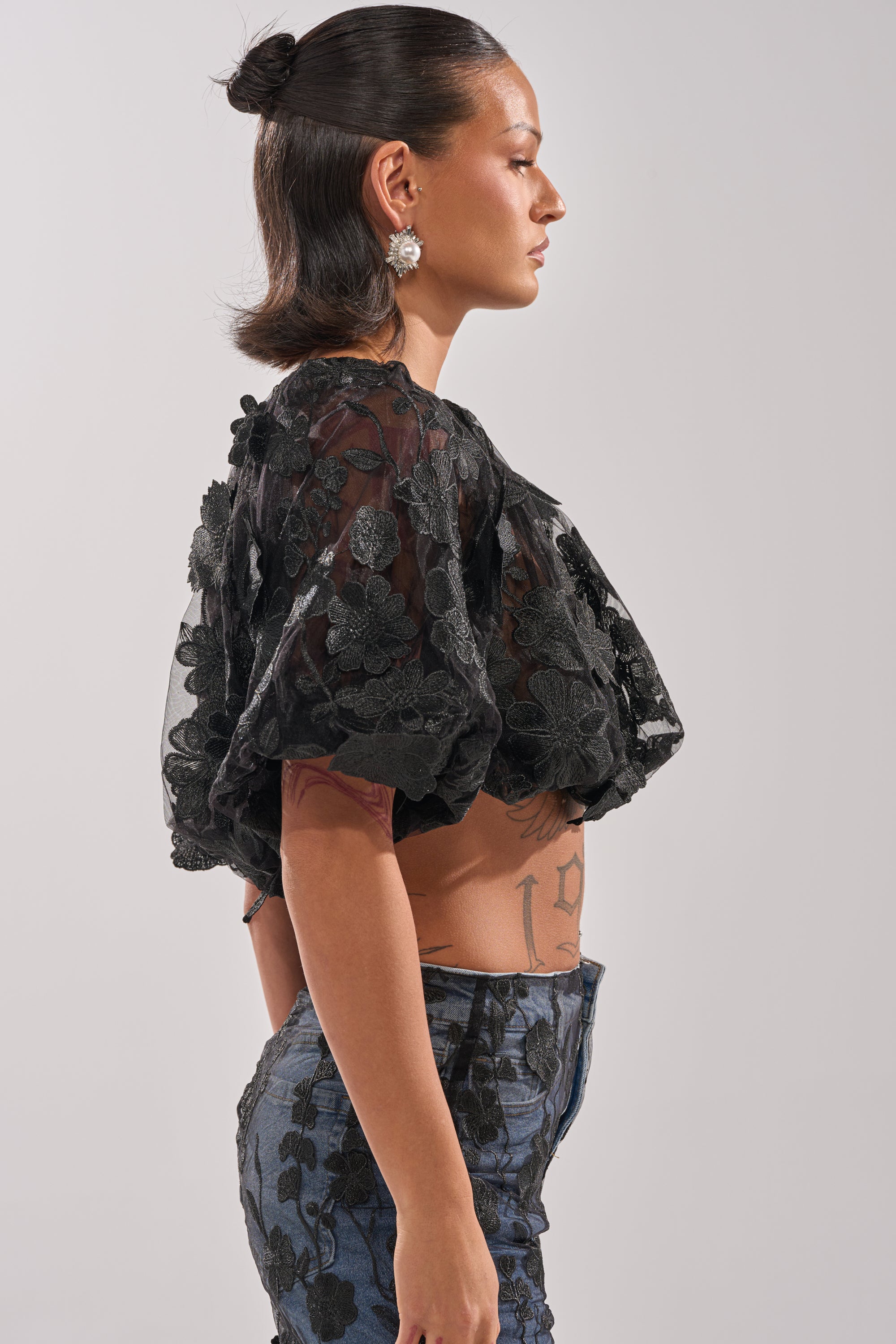 A person stands in profile wearing the DONT GIVE UP LACE BLOUSE, a sheer black floral embroidered top with puff sleeves, paired with blue high-waisted pants, large earrings, and a smooth half-up bun. Their side tattoo is visible. 2