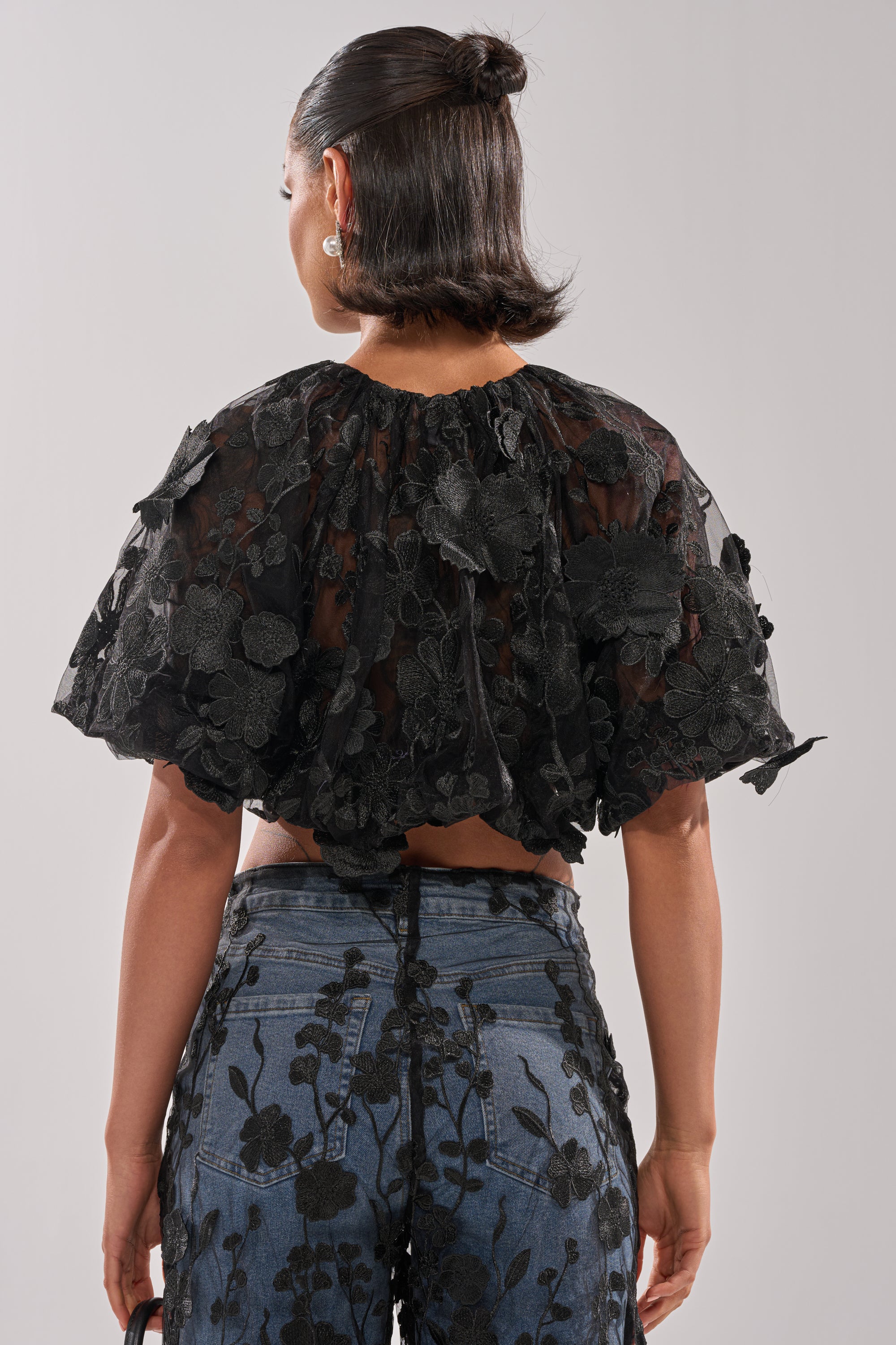A woman with dark, shoulder-length hair in a half-up bun is seen from the back wearing the DONT GIVE UP LACE BLOUSE and high-waisted blue jeans with black floral embroidery. 3