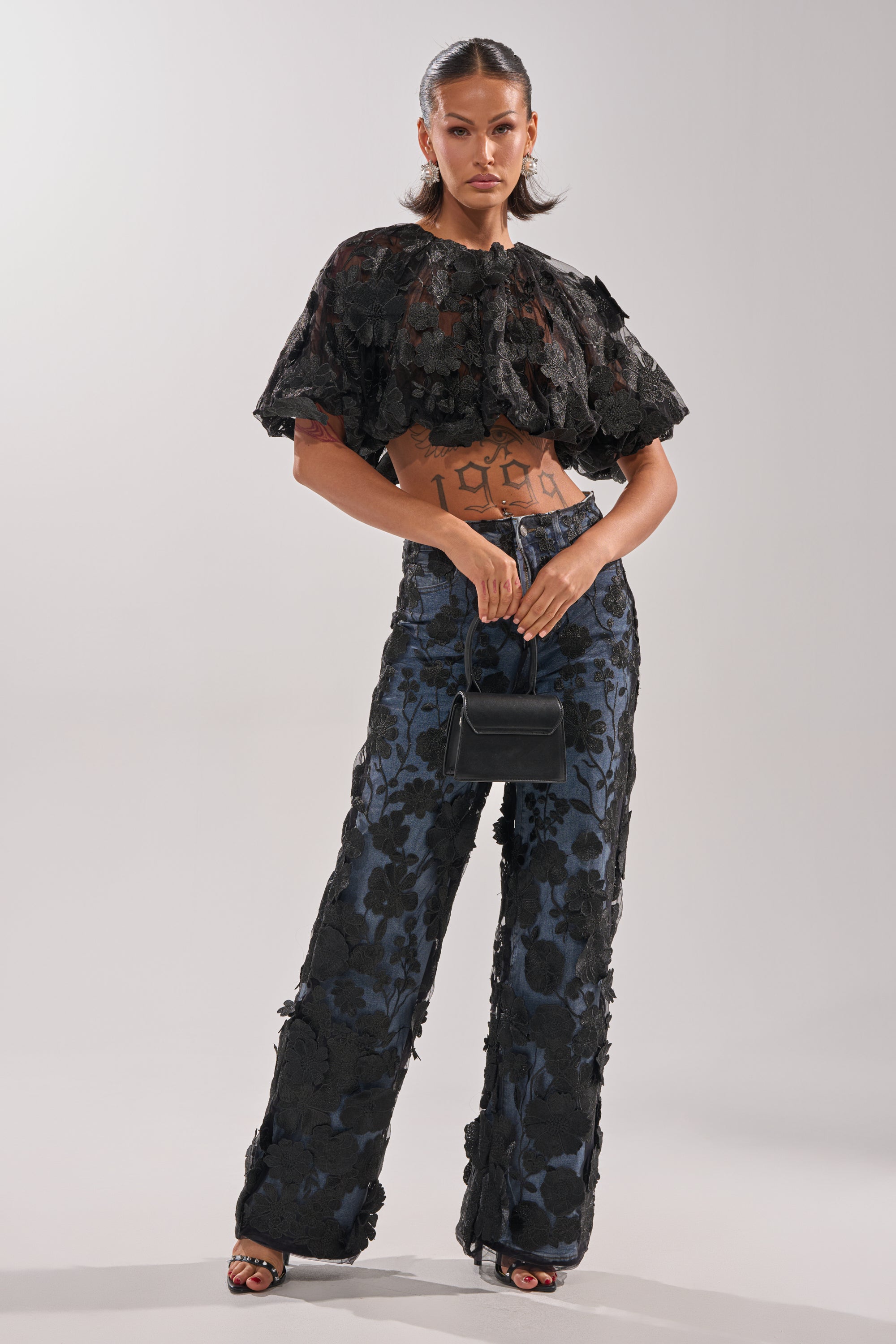 A woman stands confidently in a black textured cropped top, DONT GIVE UP RELAXED DENIM WITH TULLE OVERLAY, black heeled sandals, and a small black handbag. Her slicked-back hair and visible tattoos accent her torso.