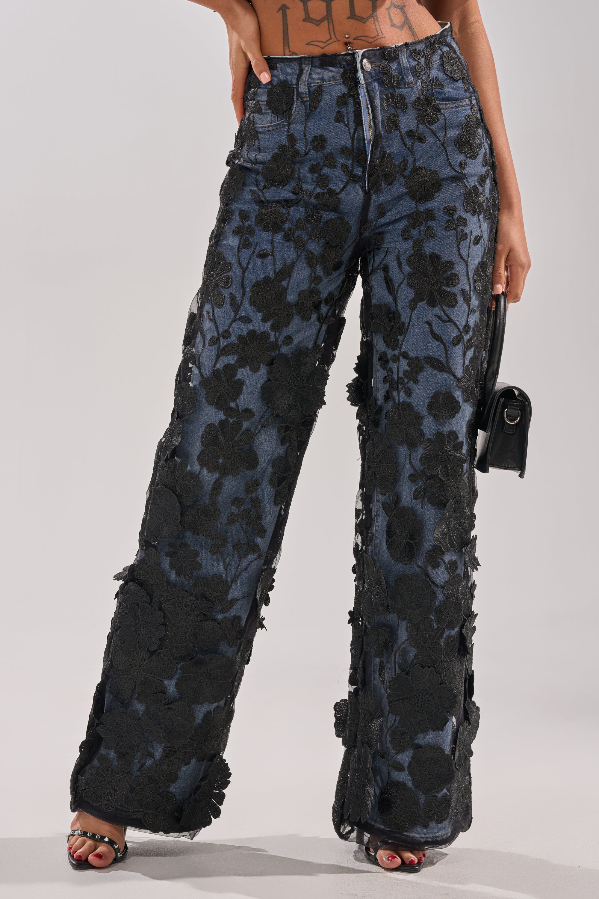 A person wears DONT GIVE UP RELAXED DENIM WITH TULLE OVERLAY: high-waisted, blue wide-leg jeans with a black tulle overlay, styled with a small black handbag. The relaxed jeans reveal their midriff and part of a tattoo.