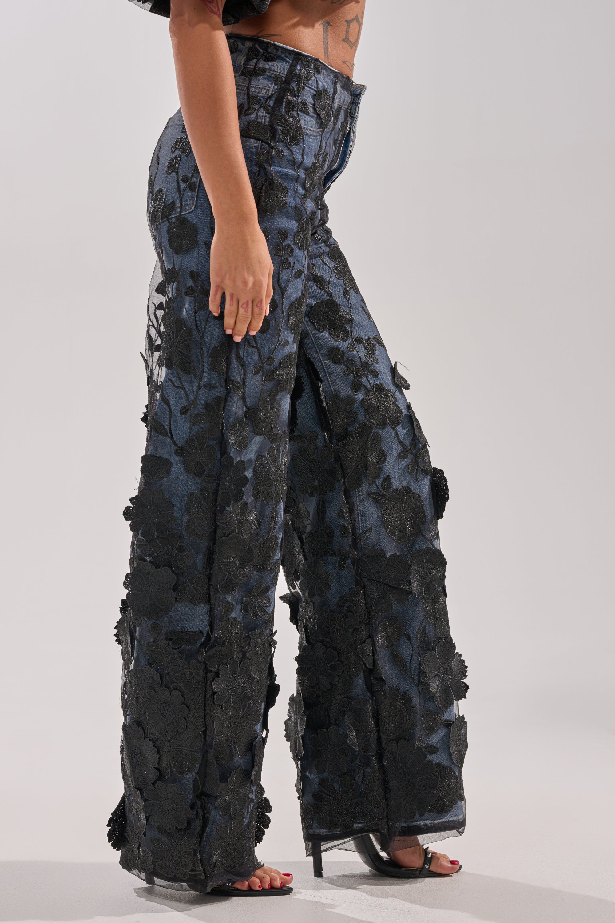 A person wears the DONT GIVE UP RELAXED DENIM WITH TULLE OVERLAY—high-waisted, wide-leg jeans featuring a tulle overlay—paired with open-toe heels, standing against a plain light background.