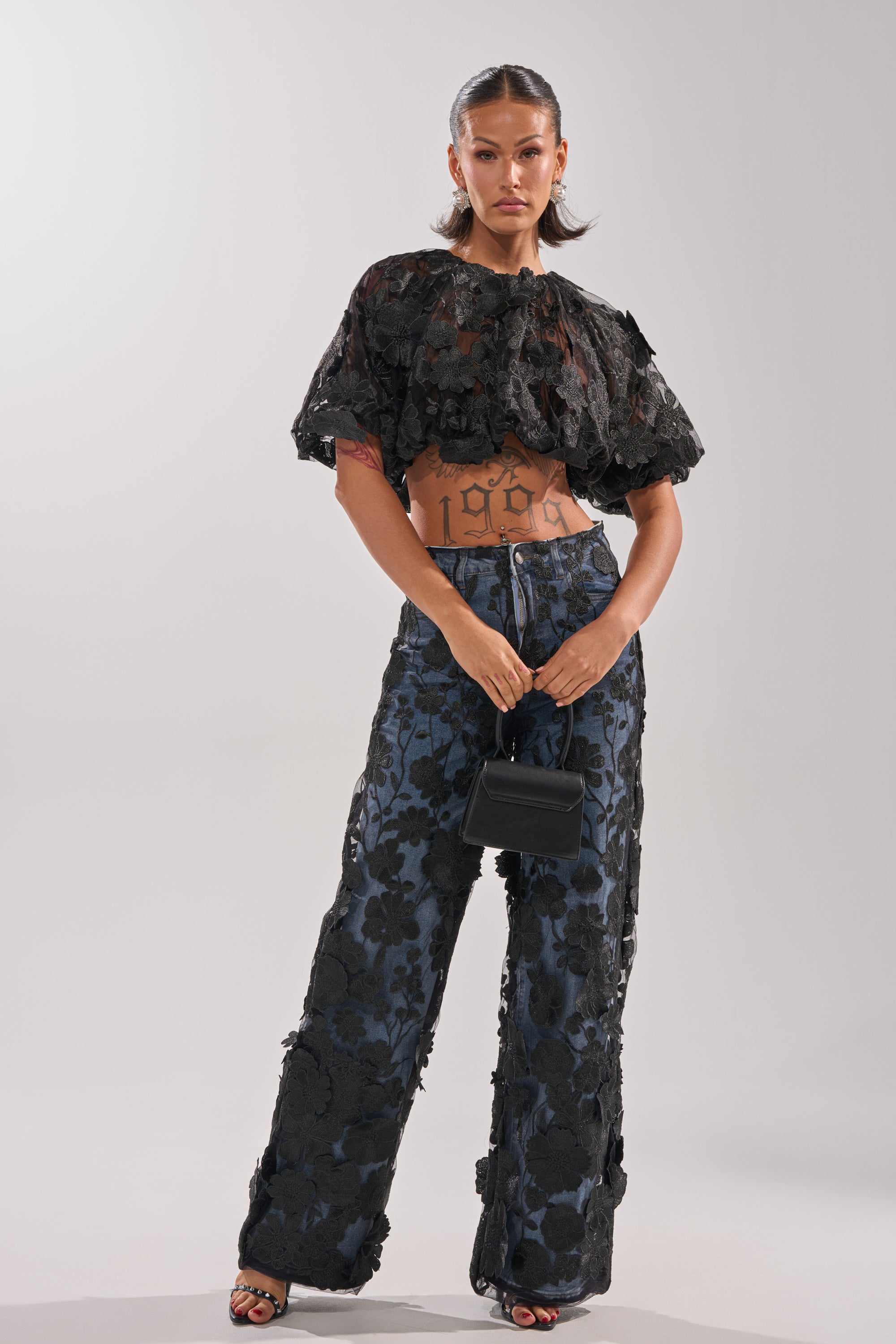 A woman stands confidently in a black textured cropped top, open-toed heels, and the DONT GIVE UP RELAXED DENIM WITH TULLE OVERLAY. She carries a small black handbag and wears her hair slicked back.