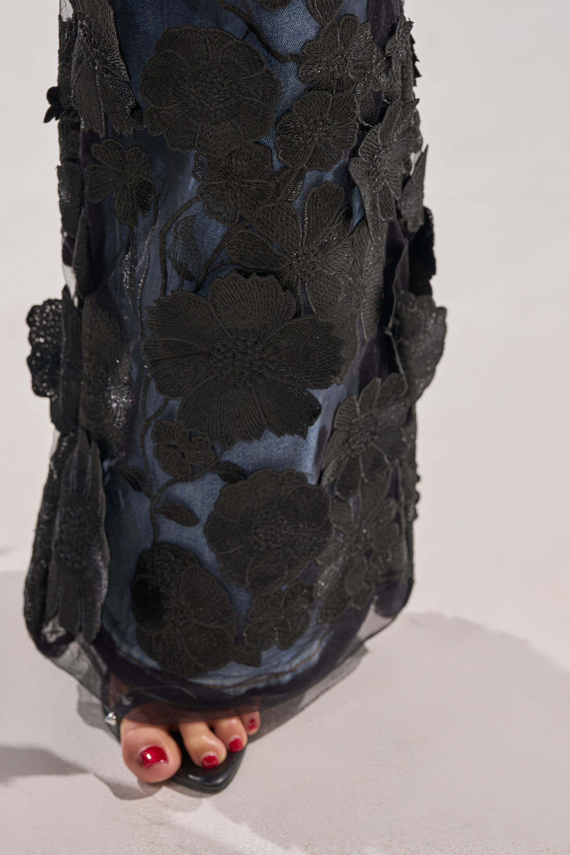 A close-up of a foot wearing the DONT GIVE UP RELAXED DENIM WITH TULLE OVERLAY, featuring sheer tulle and bold black appliqué details. The person’s toenails are painted red, shown against a soft, pale background.