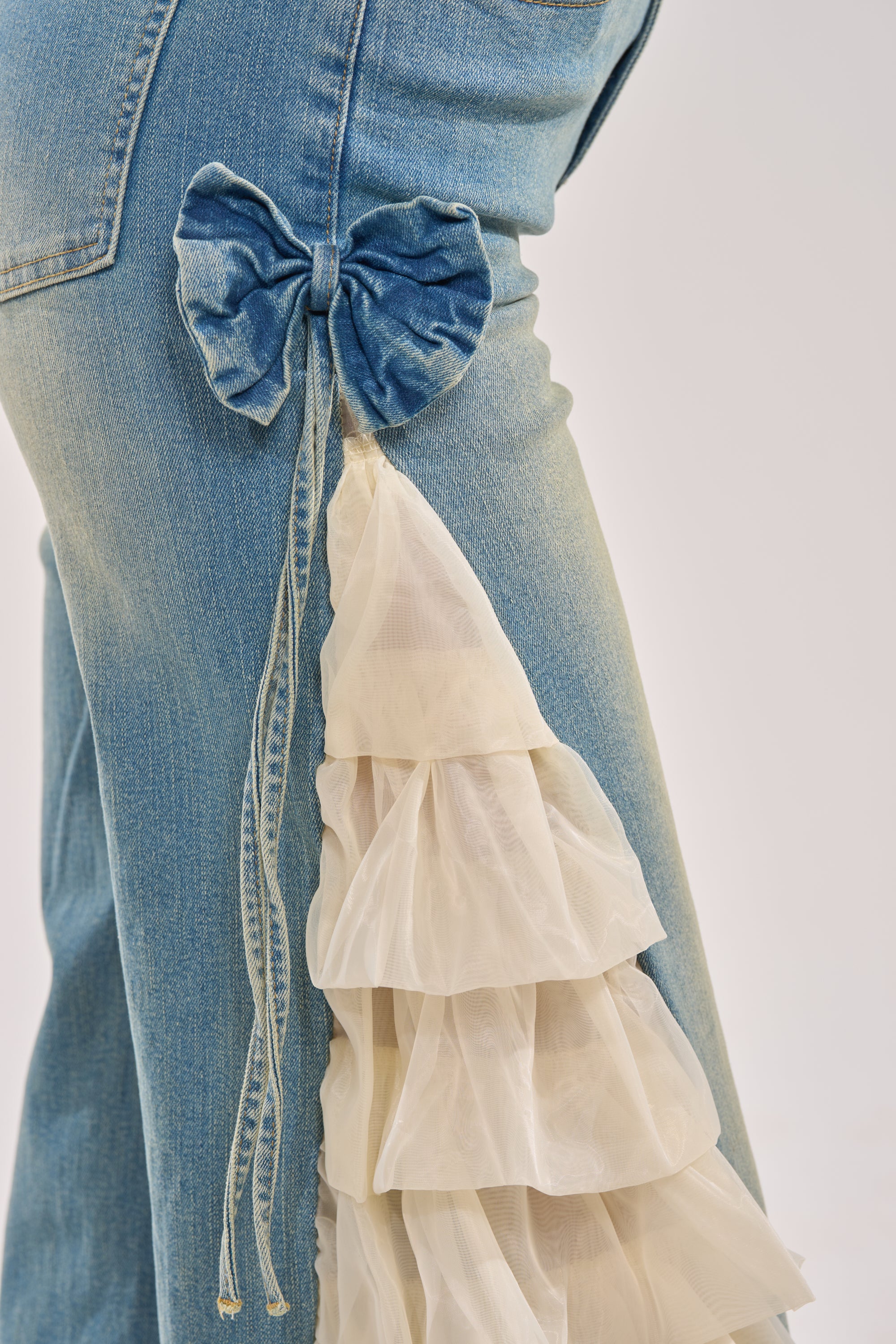 Close-up of the RUFFLE CITY STRAIGHT LEG DENIM, showcasing a decorative denim bow and layered white organza ruffle cascading from the bow for a playful, unique side detail on these straight-leg jeans. 11