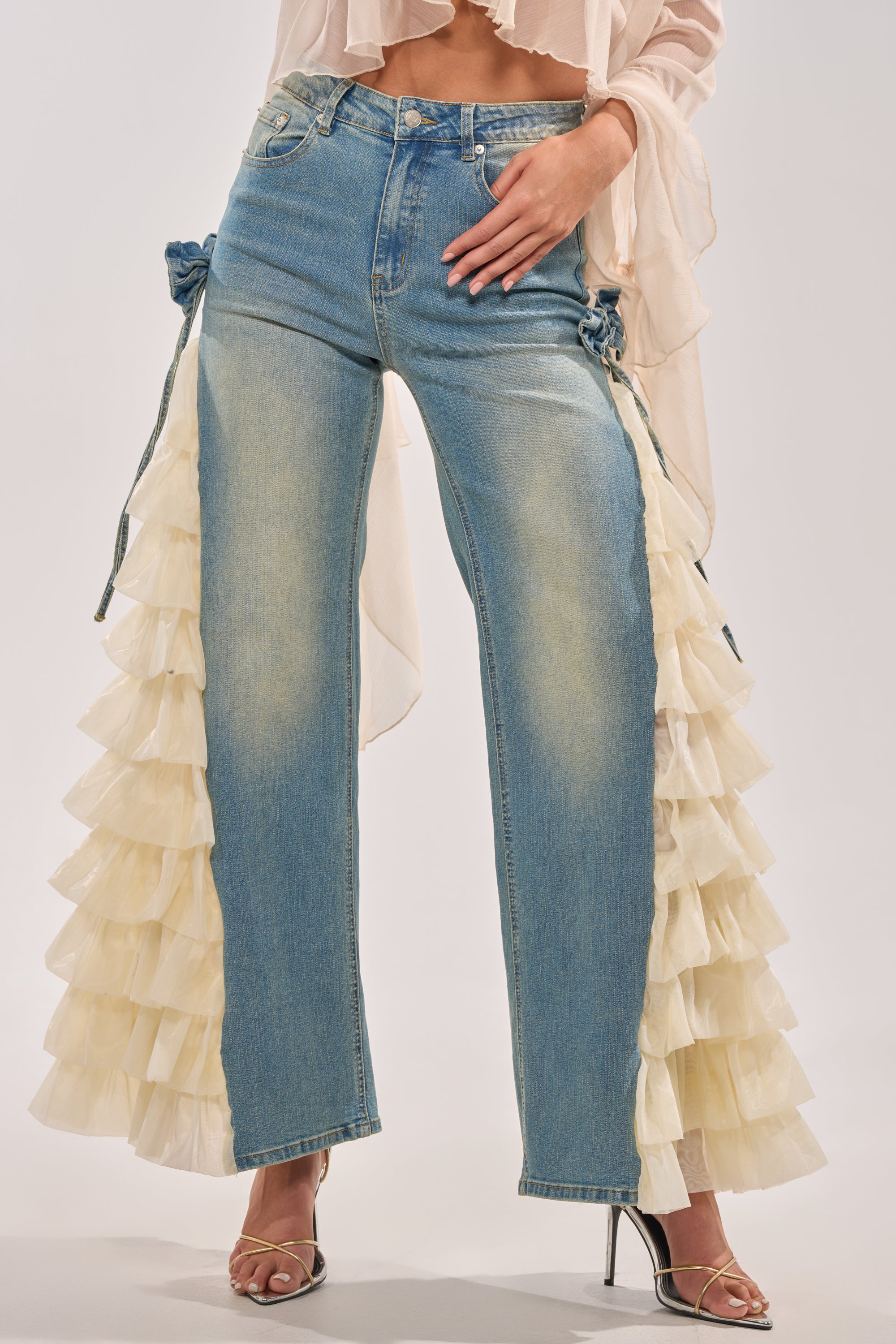 A person wears the RUFFLE CITY STRAIGHT LEG DENIM, paired with a sheer beige top and metallic open-toe heels. The background is plain and light-colored. 3