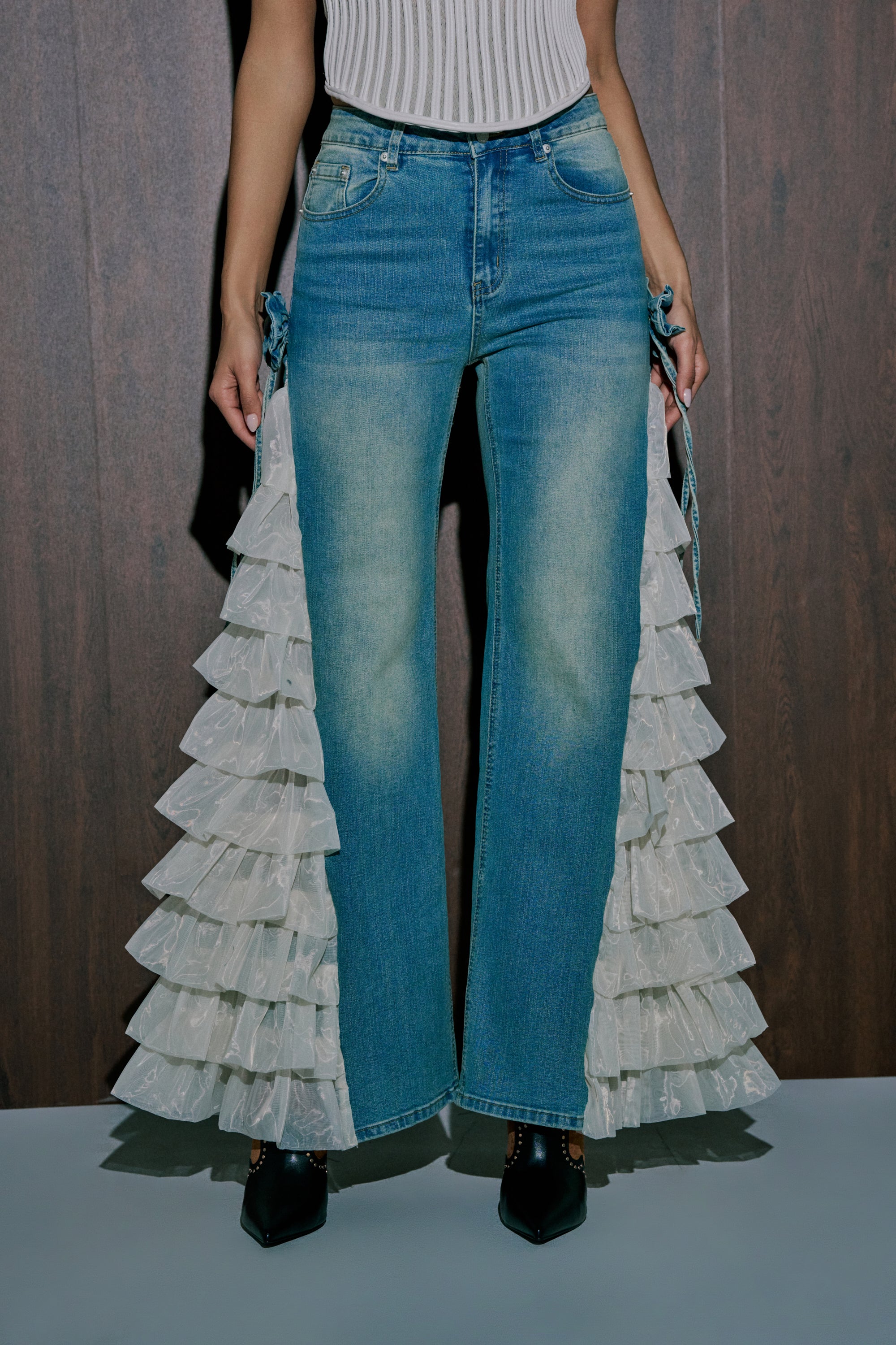 Wearing the RUFFLE CITY STRAIGHT LEG DENIM with white ruffle inserts, a person pairs them with a white top and black pointed-toe shoes, standing before a wooden background. 1