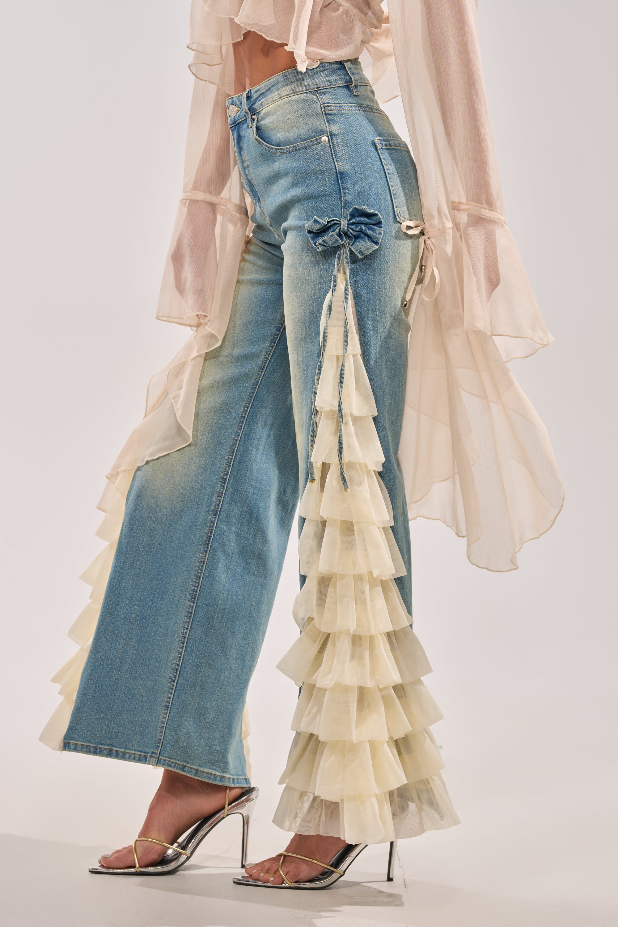Wearing the RUFFLE CITY STRAIGHT LEG DENIM featuring layered cream ruffles along the side, styled with a sheer blouse and silver high-heeled sandals for a chic, fashion-forward look. 5