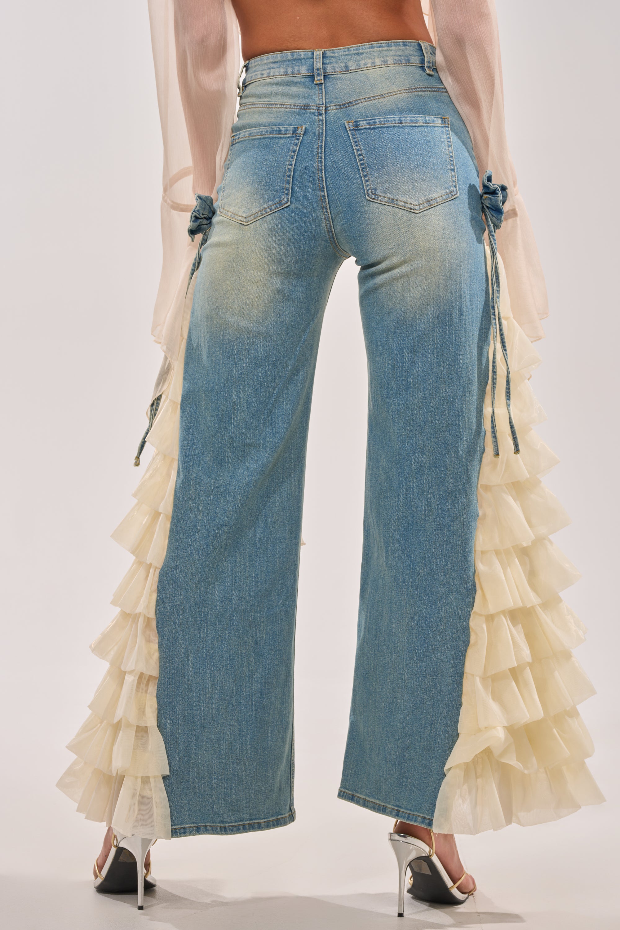 A person wears the RUFFLE CITY STRAIGHT LEG DENIM—light blue high-rise jeans with cream tulle side panels—paired with sheer long sleeves and white heels, photographed from the back. 6