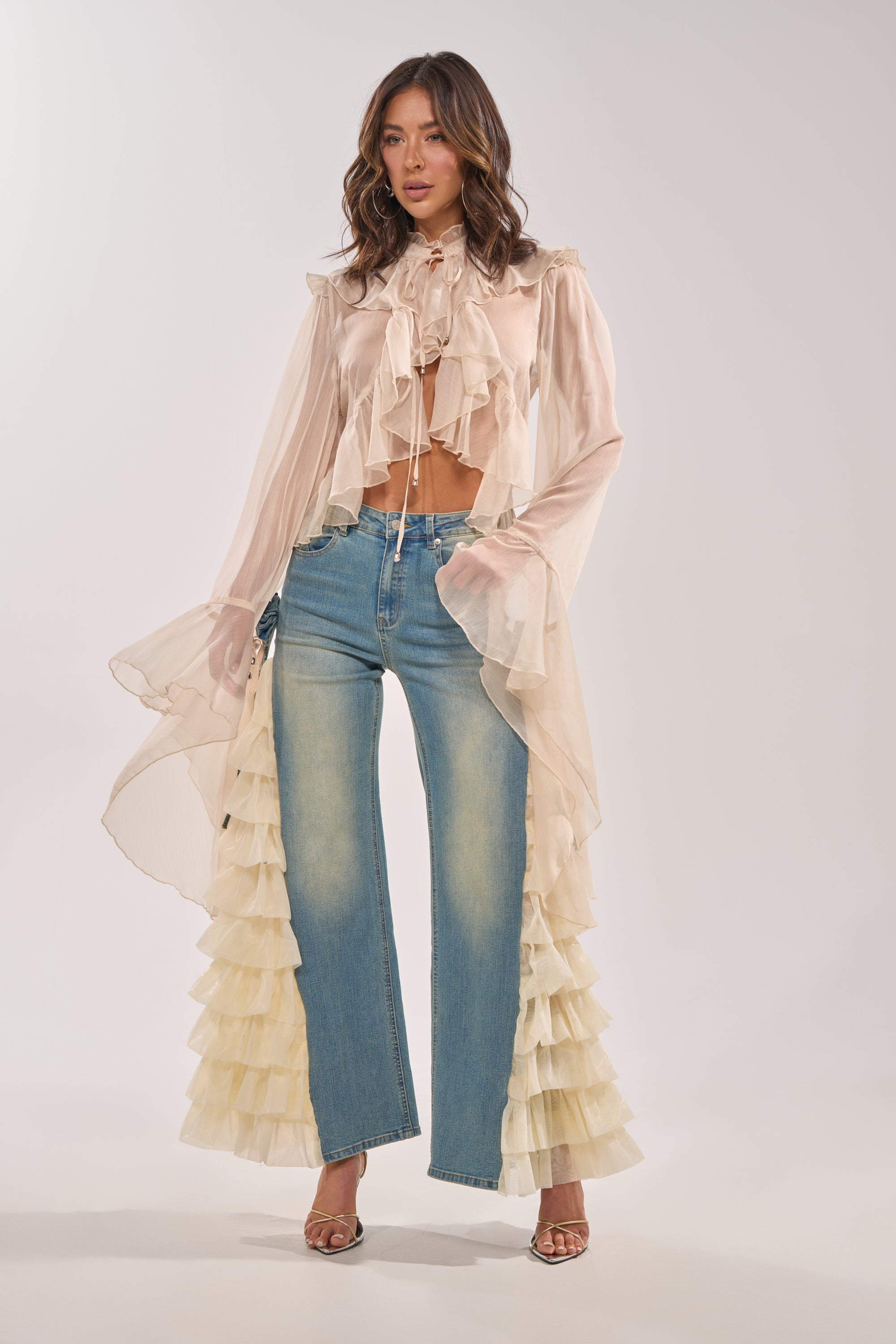 A woman models high-rise RUFFLE CITY STRAIGHT LEG DENIM with layered side ruffle details, styled with a sheer cream blouse featuring dramatic sleeves and a tie-neck, and finished with open-toe heels. 7