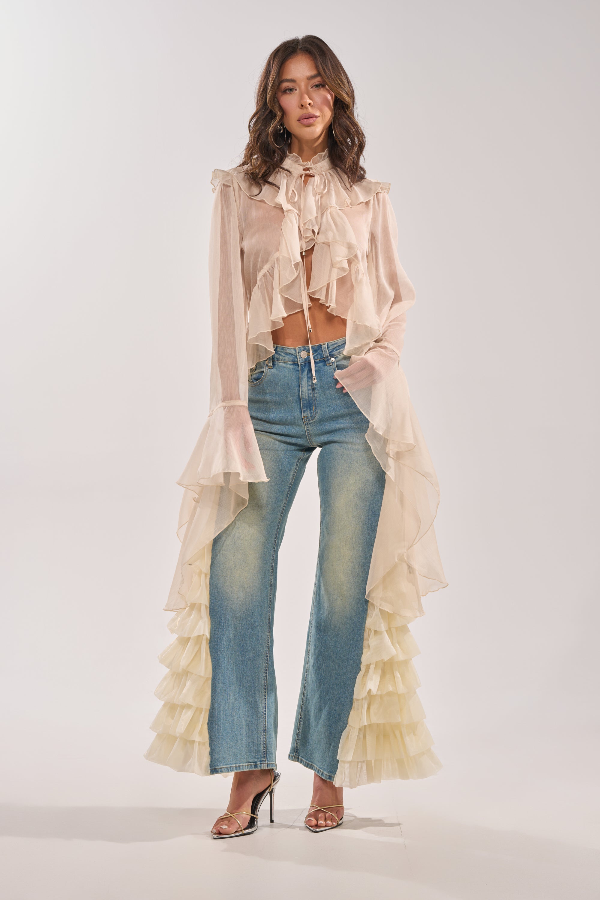 A woman wears the RUFFLE CITY STRAIGHT LEG DENIM, styled with a sheer, ruffled pale blouse featuring flared sleeves and cream side ruffles, paired with open-toe heels against a simple background. 8