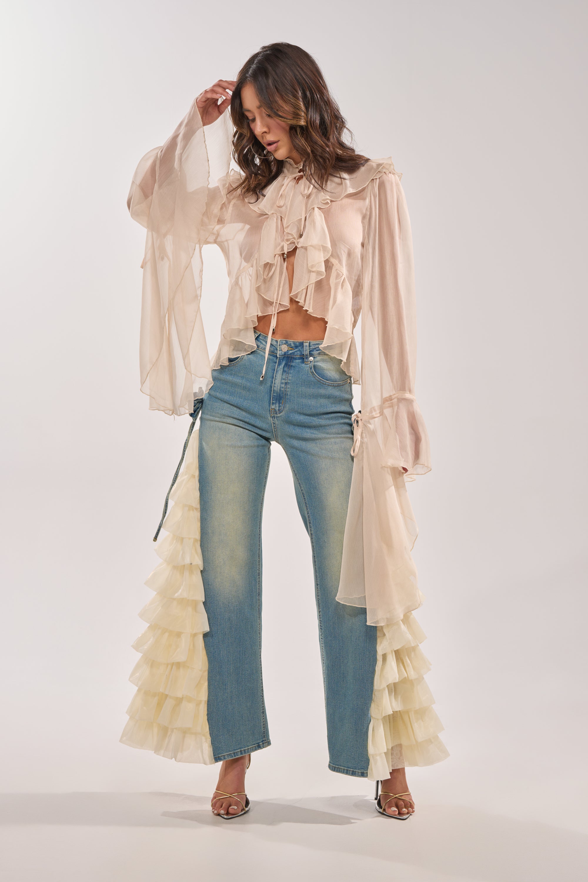 A woman poses against a plain background, wearing RUFFLE CITY STRAIGHT LEG DENIM—high-rise denim jeans with layered cream ruffles on the sides—and a sheer, ruffled beige blouse, with one hand touching her hair. 9