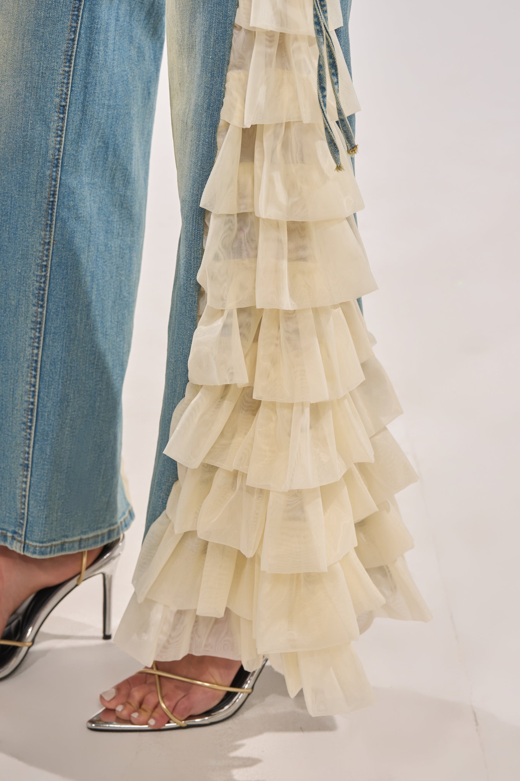A close-up of a person wearing RUFFLE CITY STRAIGHT LEG DENIM, featuring blue jeans with layered cream-colored organza ruffles, paired with silver pointed-toe heels with thin ankle straps, standing on a light surface. 10