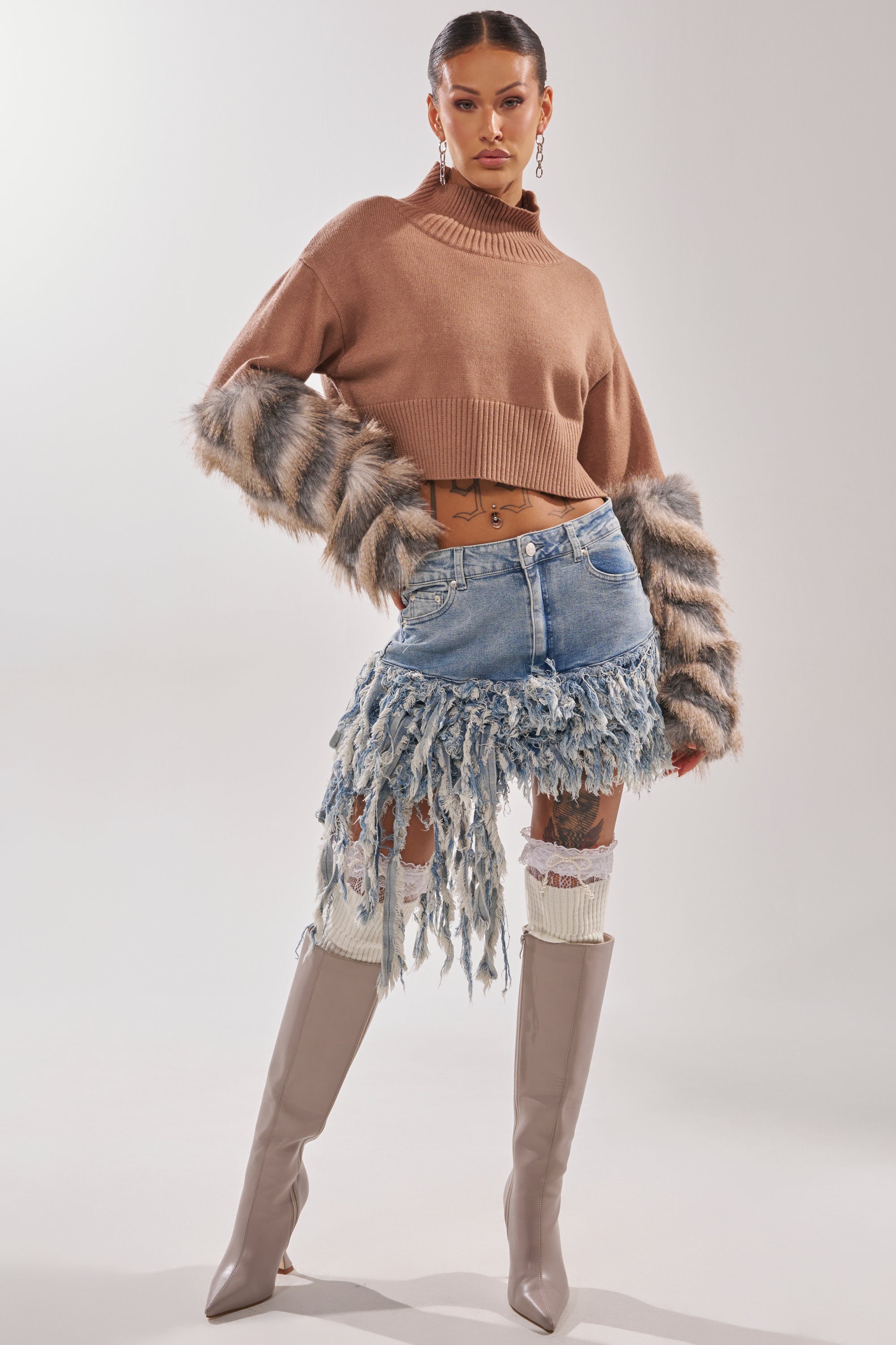 A woman poses in the OPEN UP YOUR LOVE DENIM SKIRT, a cropped brown sweater with furry sleeves, white leg warmers, and knee-high taupe boots against a plain background. 2