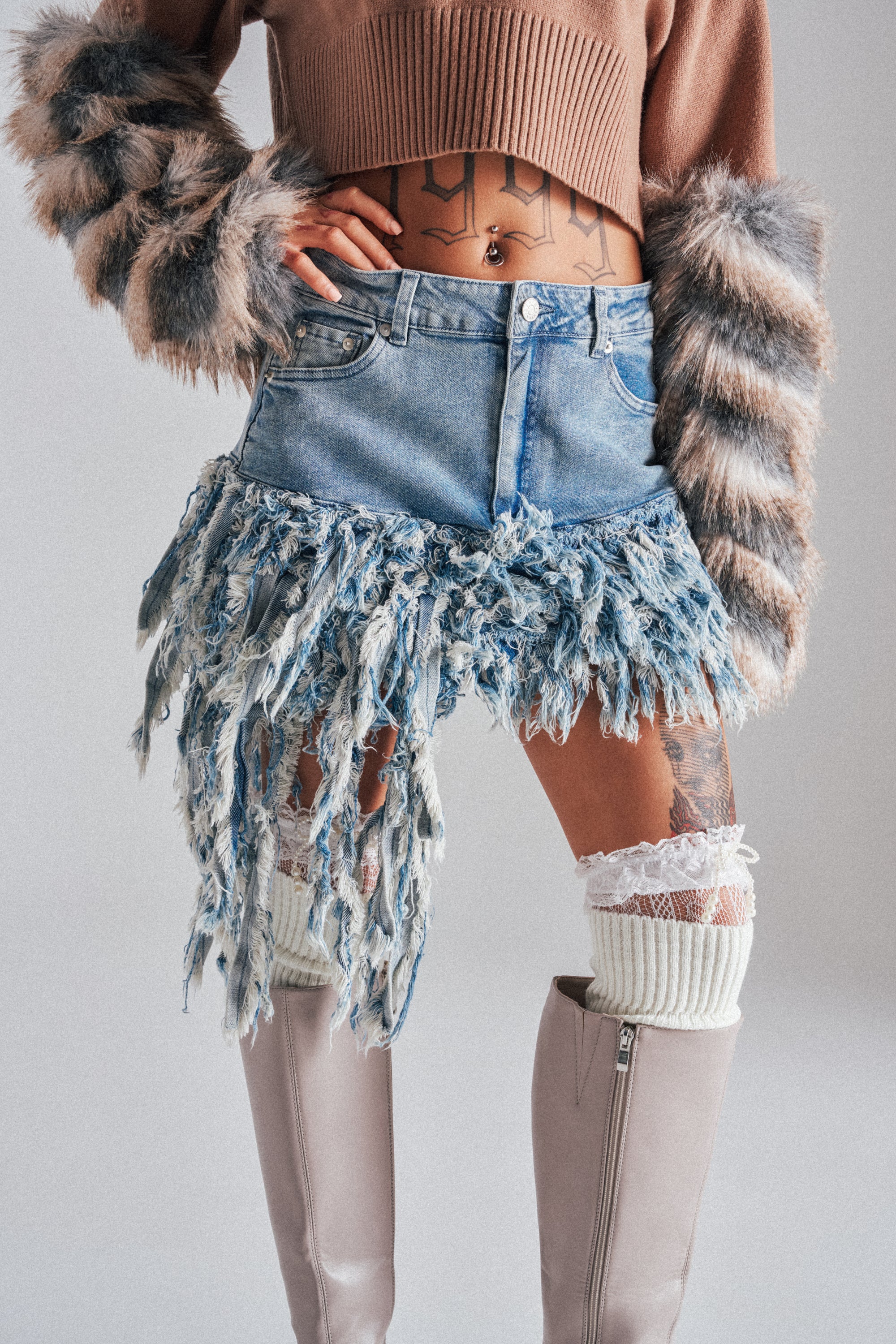 A person wearing a cropped brown sweater with furry sleeves, the OPEN UP YOUR LOVE DENIM SKIRT—a distressed mini skirt with frayed denim strips—beige knee-high boots, and white thigh-high socks with lace accents. 0