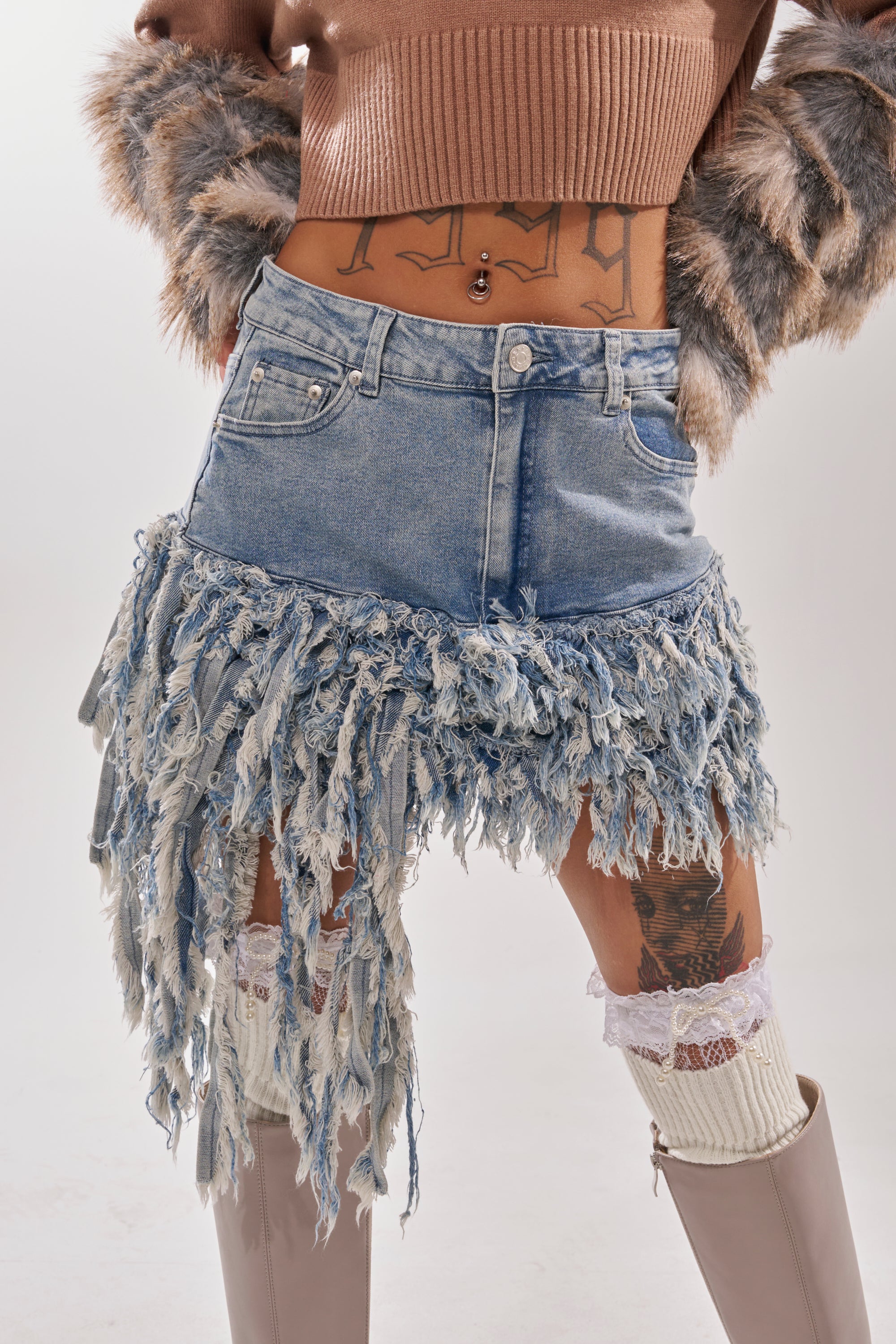 A person wearing a cropped tan sweater with faux fur sleeves, the OPEN UP YOUR LOVE DENIM SKIRT featuring long frayed tassels, thigh tattoos, white lace garters, knitted leg warmers, and beige knee-high boots. 3