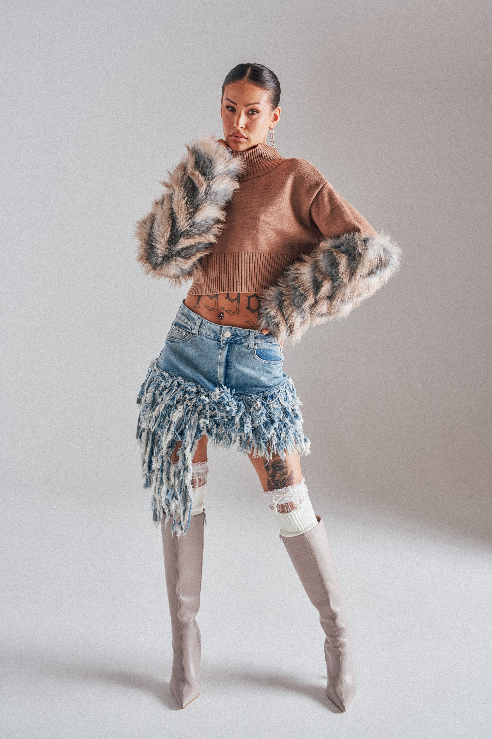 A person poses in the OPEN UP YOUR LOVE DENIM SKIRT paired with a tan cropped sweater with faux fur sleeves, white knee-high socks, and tall beige boots, standing against a neutral background. 1