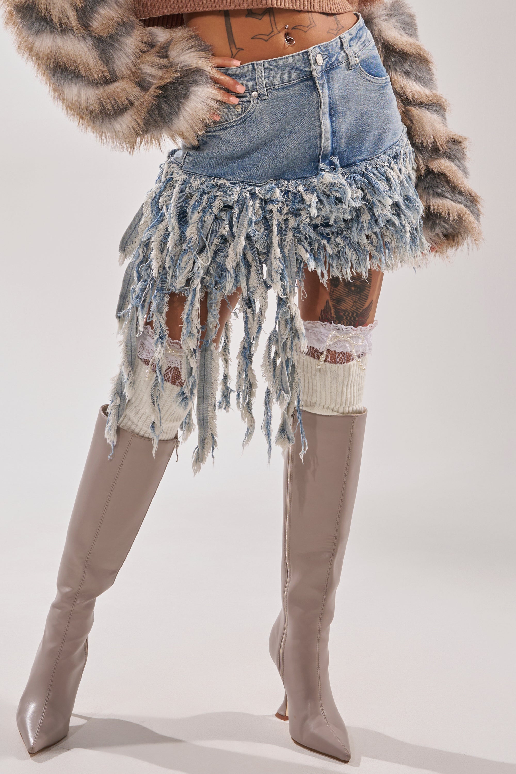 A person is wearing the OPEN UP YOUR LOVE DENIM SKIRT with long shredded fringes, a cropped fur jacket, lace-trimmed leg warmers, taupe knee-high boots, and has visible thigh tattoos and a belly button piercing. 4
