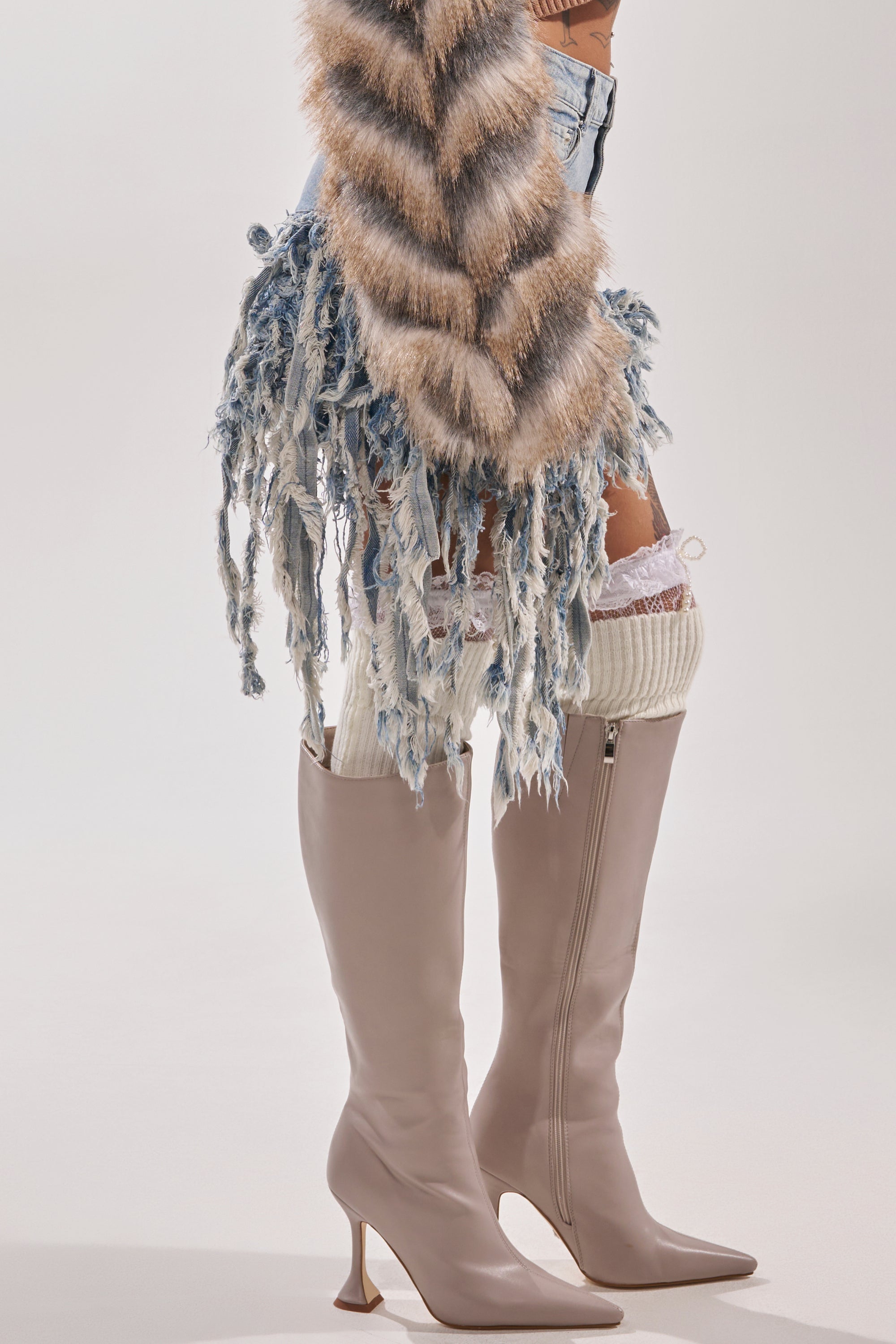 A person poses against a light background wearing tall beige heeled boots, white knit leg warmers, a brown-and-cream faux fur garment, and the OPEN UP YOUR LOVE DENIM SKIRT. 5