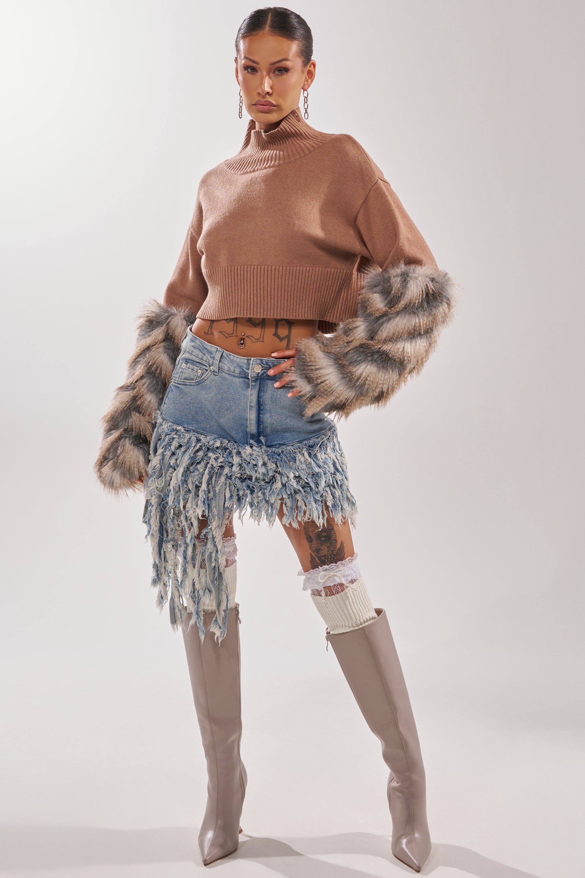 A woman stands confidently in the OPEN UP YOUR LOVE DENIM SKIRT, styled with a brown cropped sweater featuring faux fur sleeves, knee-high beige boots, white socks, and hoop earrings against a plain background. 8