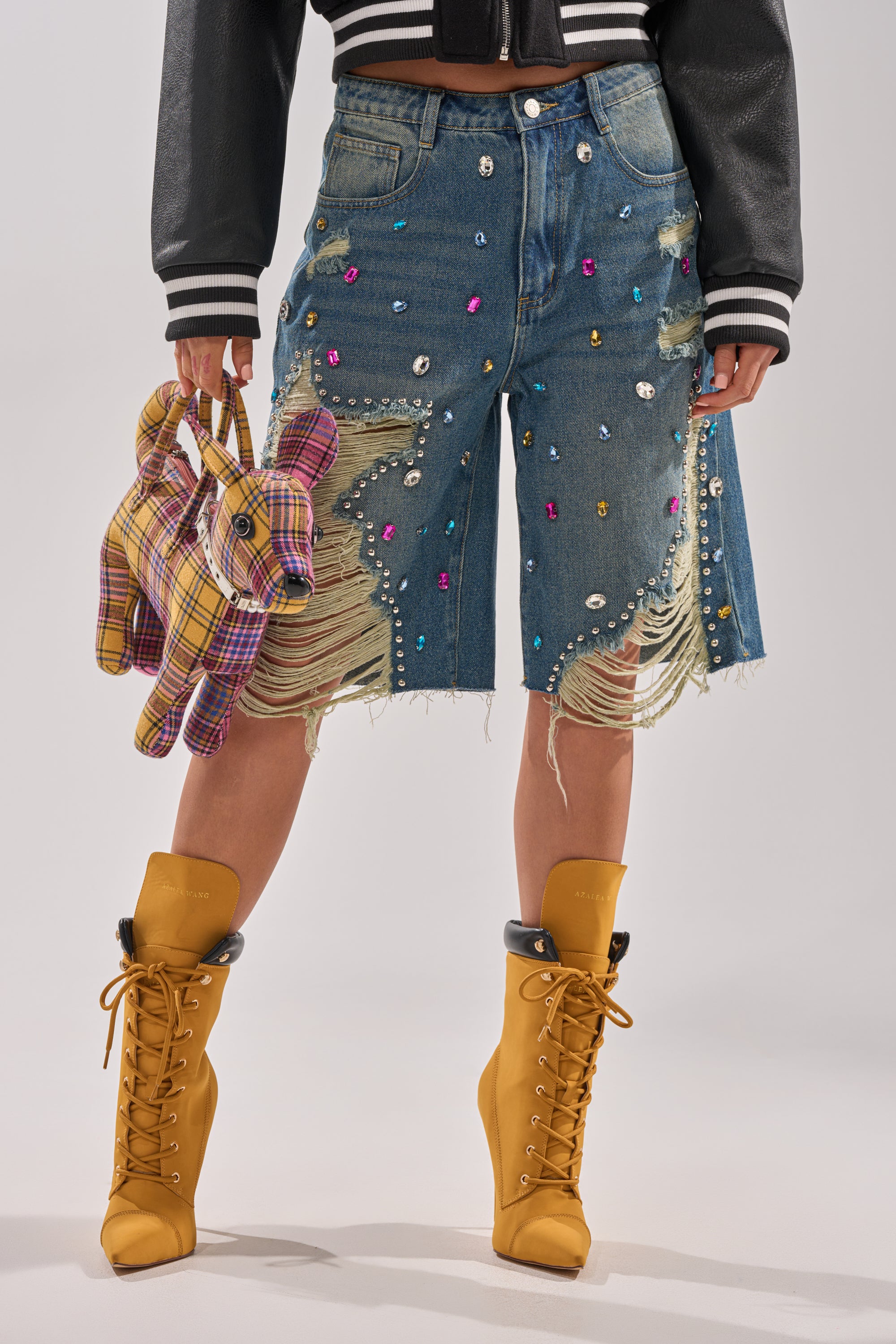 A person wears the JEWELS FOR DAYS BERMUDA SHORT, embellished with colorful gems, paired with tan lace-up heeled boots, a plaid dog-shaped handbag, and a black cropped jacket. 0
