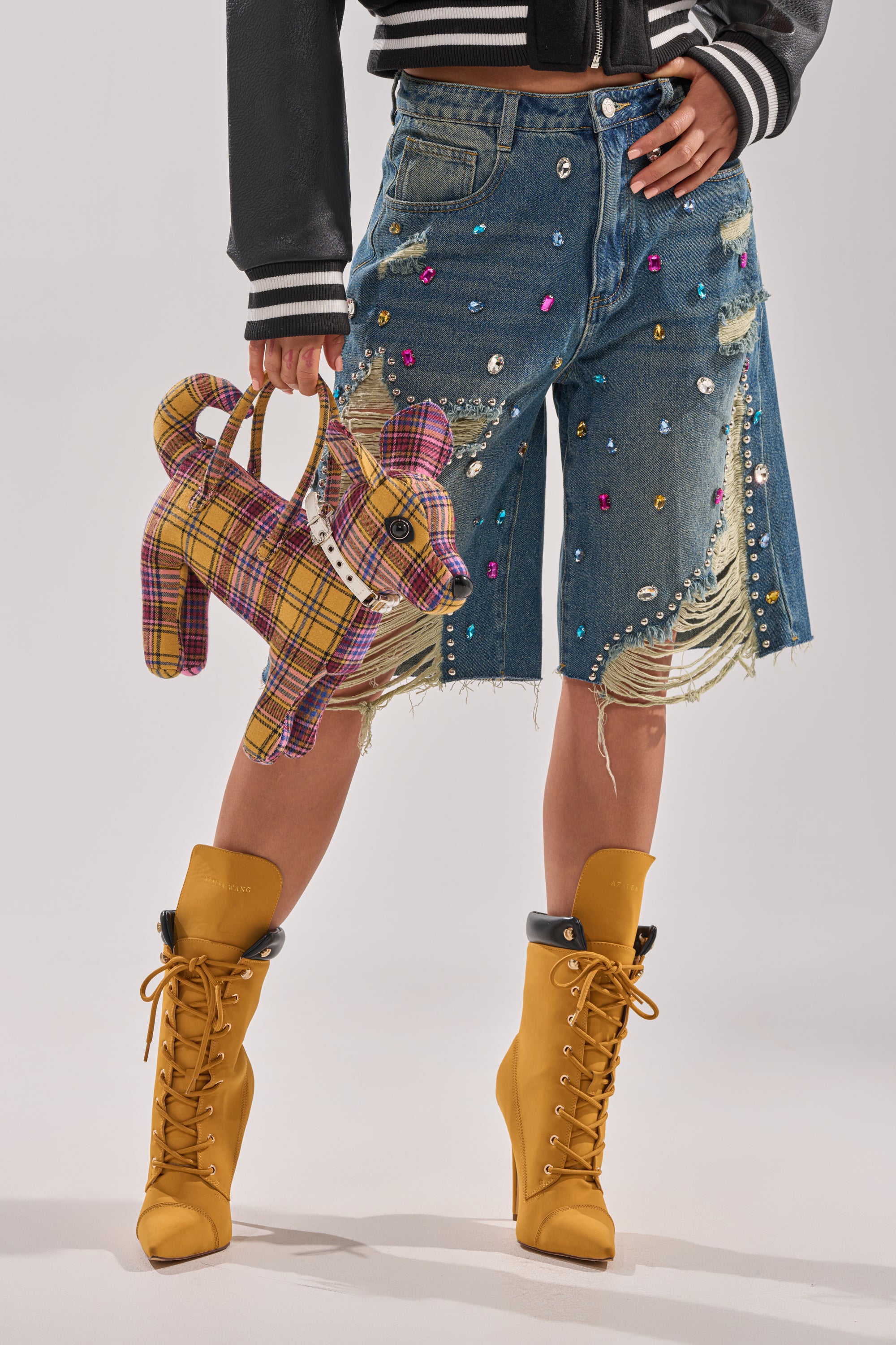 Wearing JEWELS FOR DAYS BERMUDA SHORTS, gemstone-embellished and distressed, tan lace-up boots, and a black-and-white jacket, the person holds a plaid dog-shaped handbag against a light plain background. 1