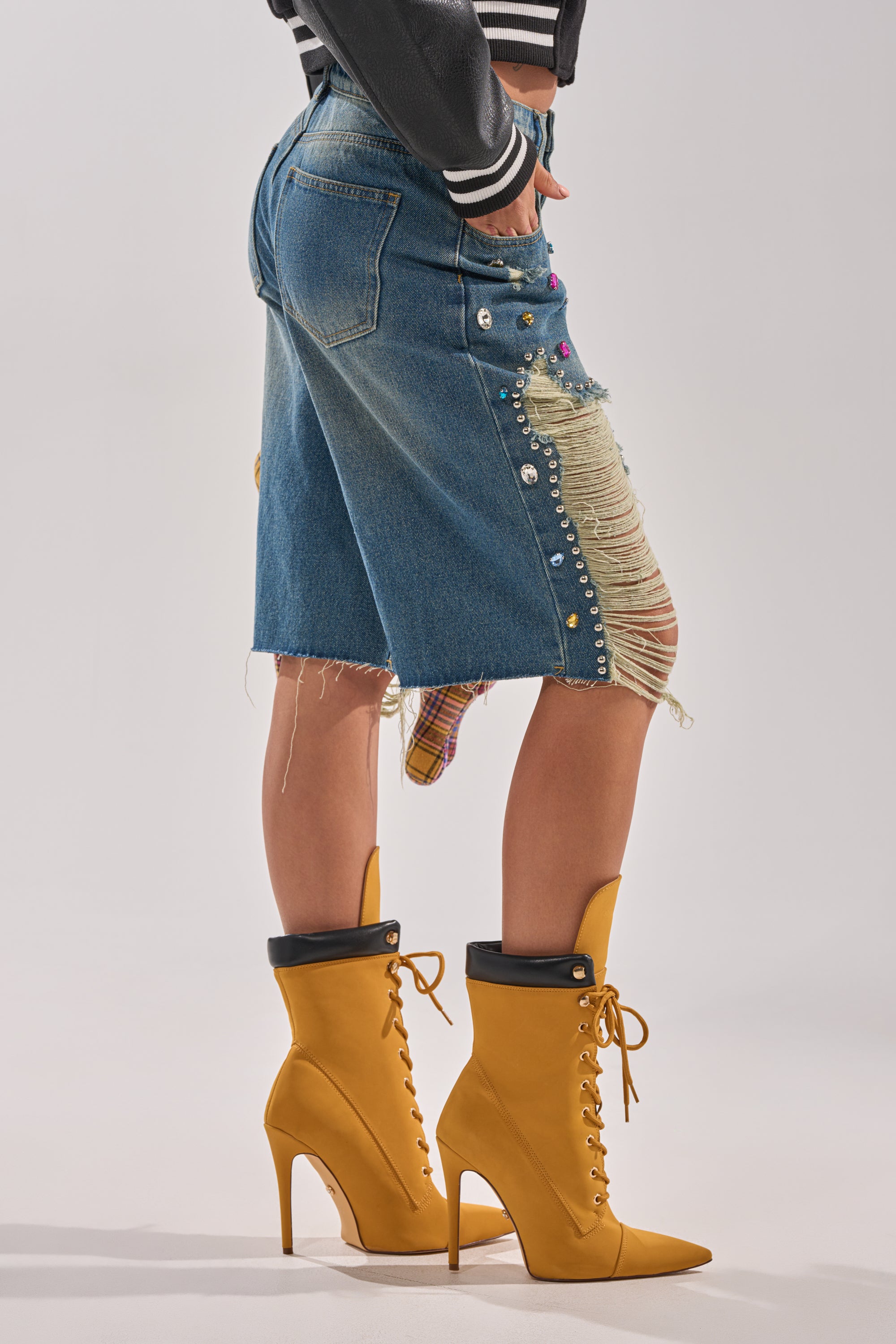 A person wearing the JEWELS FOR DAYS BERMUDA SHORT, featuring decorative studs and plaid patches, paired with mustard yellow high-heeled lace-up boots. Gemstone embellishments add sparkle against a plain, light background. 2