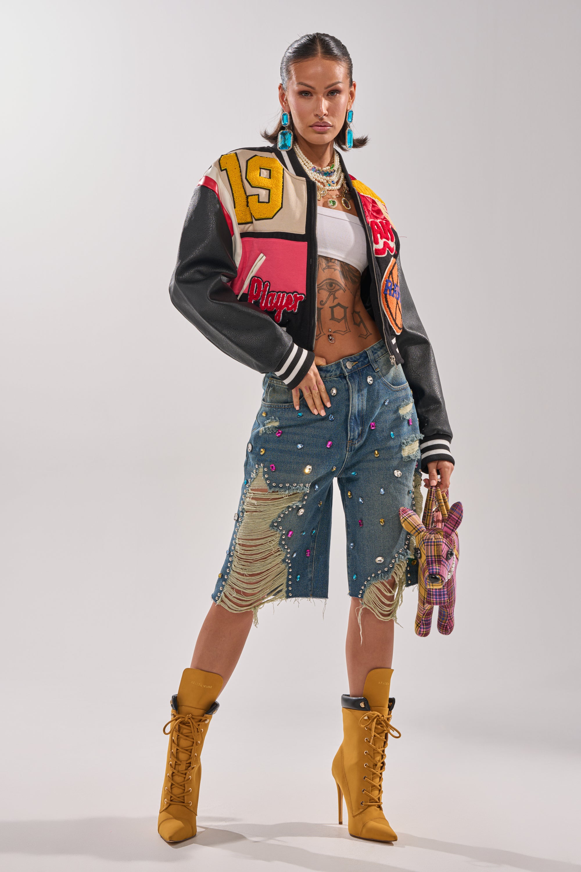 A woman with slicked-back hair and tattoos poses confidently in the JEWELS FOR DAYS BERMUDA SHORT, styled with a varsity jacket, white crop top, tan heeled boots, statement earrings, and a plaid stuffed animal. 5