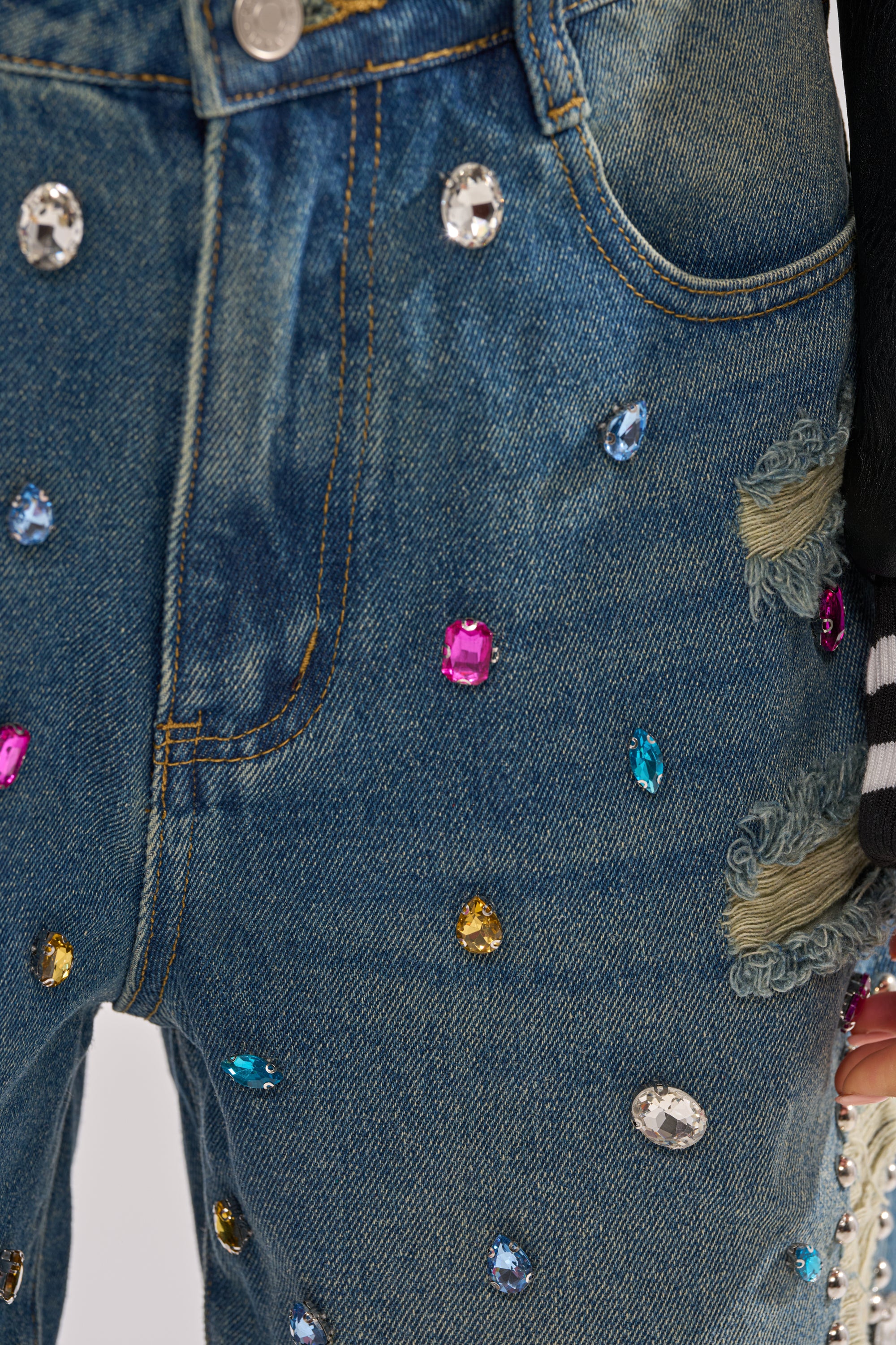 Close-up of the JEWELS FOR DAYS BERMUDA SHORT, featuring distressed denim with colorful gemstone embellishments, frayed patches, and a silver waistband button. A black and white striped sleeve appears on the right. 6