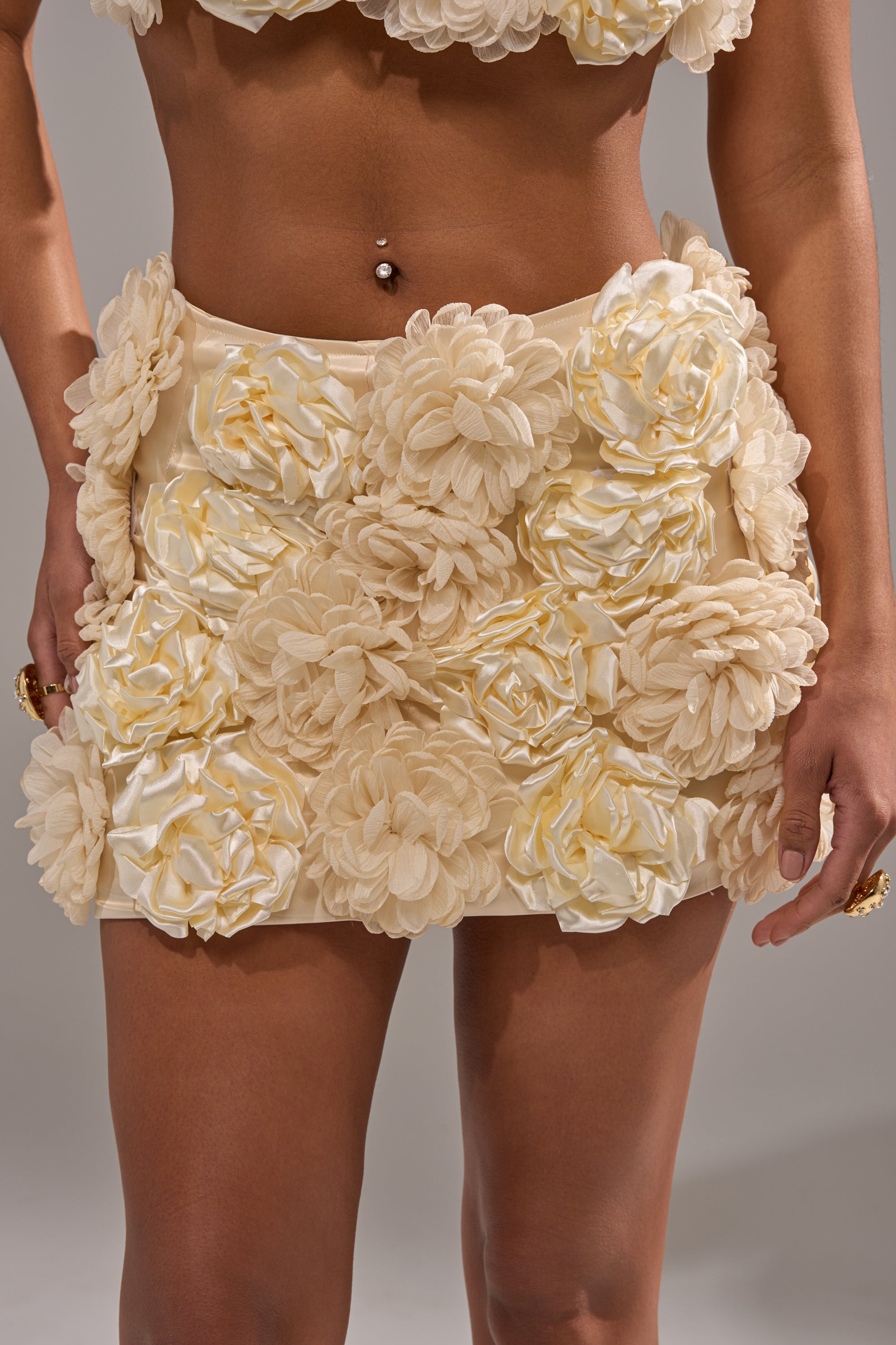 Someone wears the FLOWER POWER MINI SKIRT, a satin high-rise mini skirt embellished with oversized textured floral appliqués, revealing their midriff and belly button piercing against a plain background.