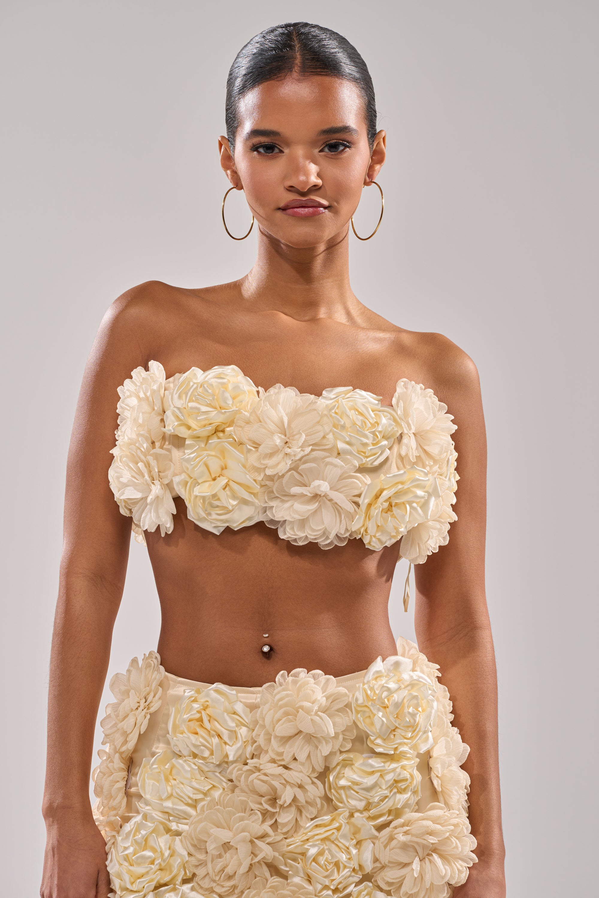 A woman with hoop earrings wears the FLOWER POWER BRALETTE, a cream-colored satin crop top with large 3D floral appliqués, paired with a matching skirt, and stands against a plain light gray background. 4