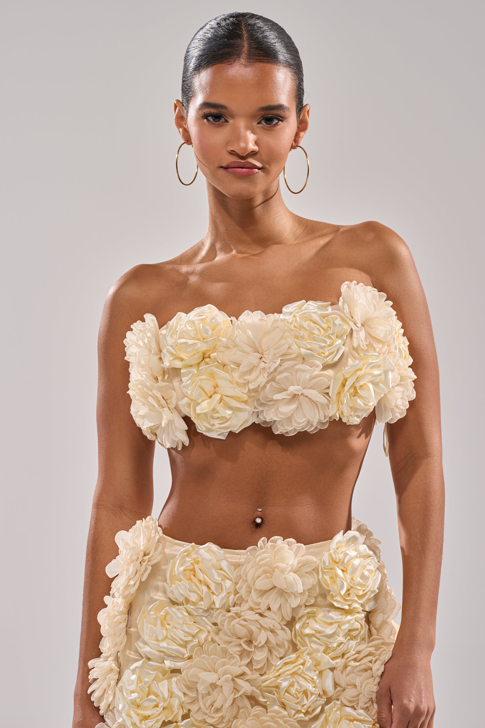 A woman models the FLOWER POWER BRALETTE, a cream strapless top with bold 3D floral appliques, styled with large gold hoop earrings and a matching skirt against a plain light background. 5