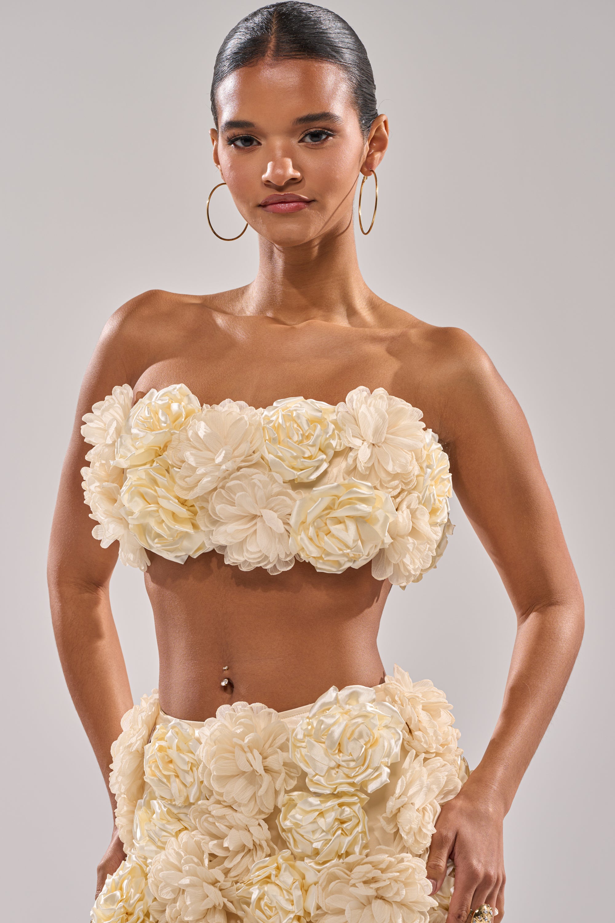 A woman with dark hair pulled back wears large hoop earrings and the FLOWER POWER BRALETTE, a cream satin style with 3D floral appliques. She stands confidently with hands on hips against a plain background. 6