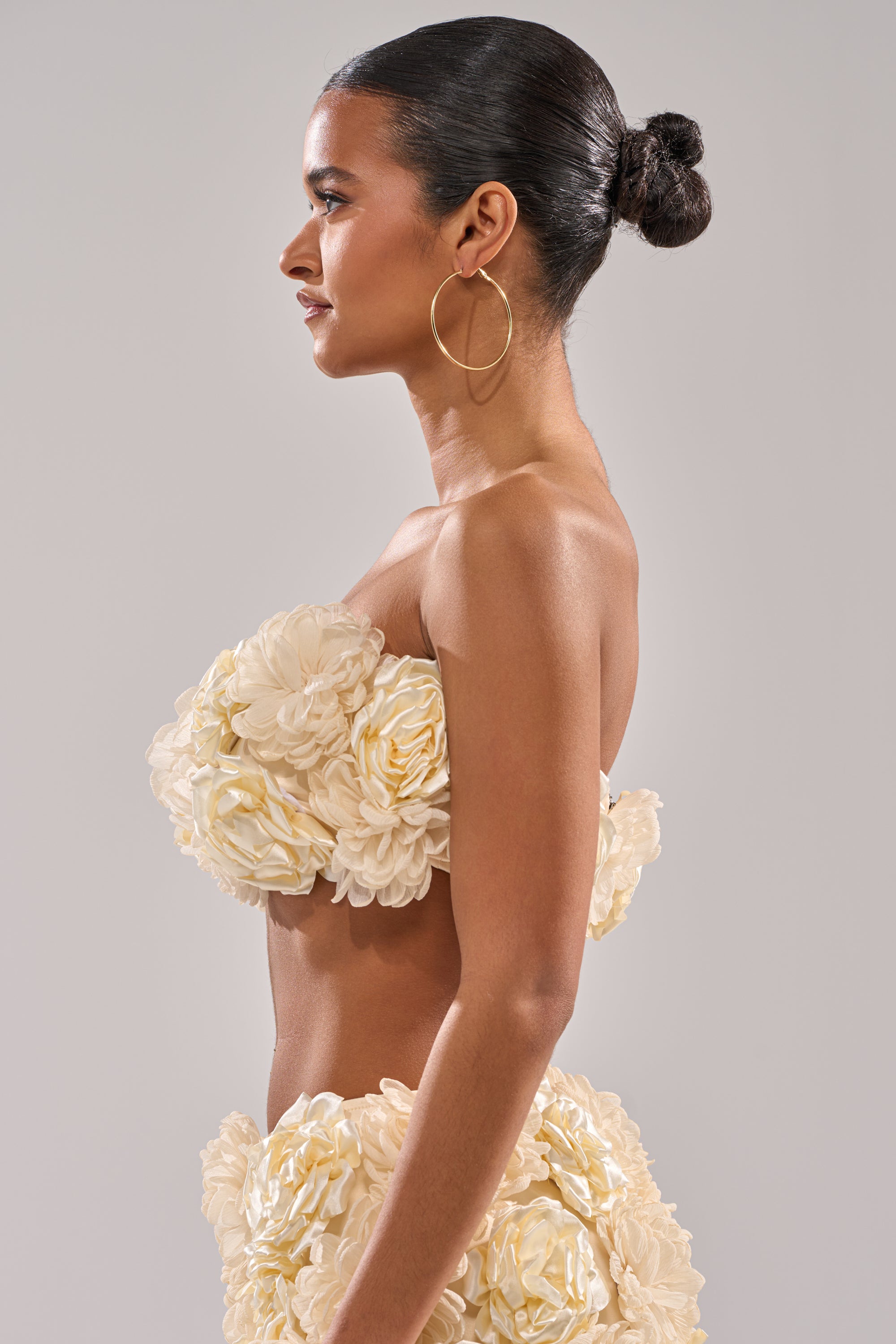 A woman with dark hair in a sleek bun, wearing large hoop earrings and the cream FLOWER POWER BRALETTE with textured, flower-like ruffles, poses in profile against a plain light gray background. 7
